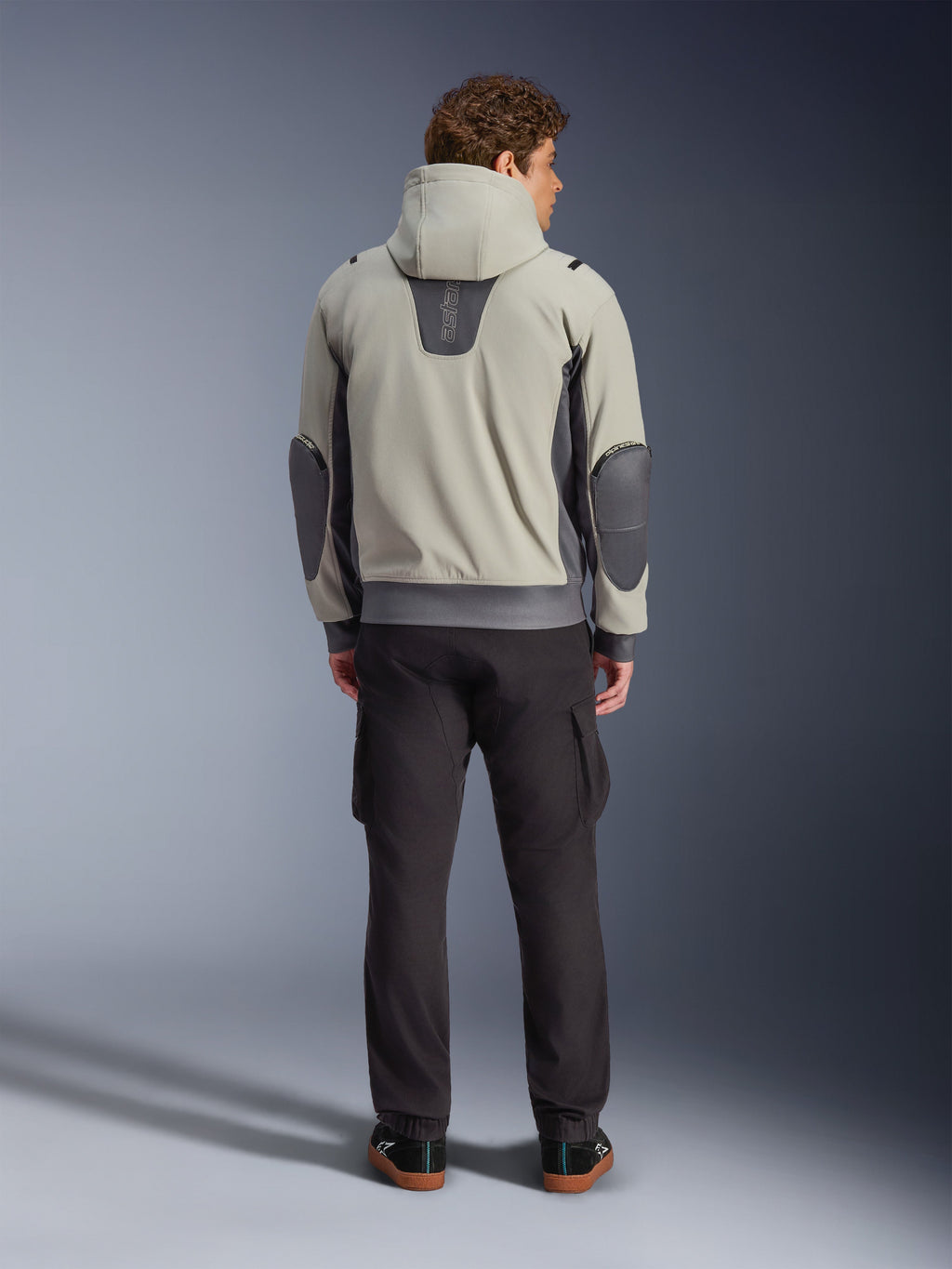 Alpinestars Moblast Waterproof Jacket, Iron Gray Dark Sage, vista posteriore di un motociclista che indossa la giacca da moto con cappuccio, colorazione grigio chiaro e verde salvia scuro, logo 'astars' verticale sulla parte superiore della schiena, toppe sui gomiti grigio scuro, abbinata a pantaloni cargo scuri per un look urbano contemporaneo.