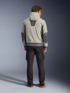 Alpinestars Moblast Waterproof Jacket, Iron Gray Dark Sage, vista posteriore di un motociclista che indossa la giacca da moto con cappuccio, colorazione grigio chiaro e verde salvia scuro, logo 'astars' verticale sulla parte superiore della schiena, toppe sui gomiti grigio scuro, abbinata a pantaloni cargo scuri per un look urbano contemporaneo.