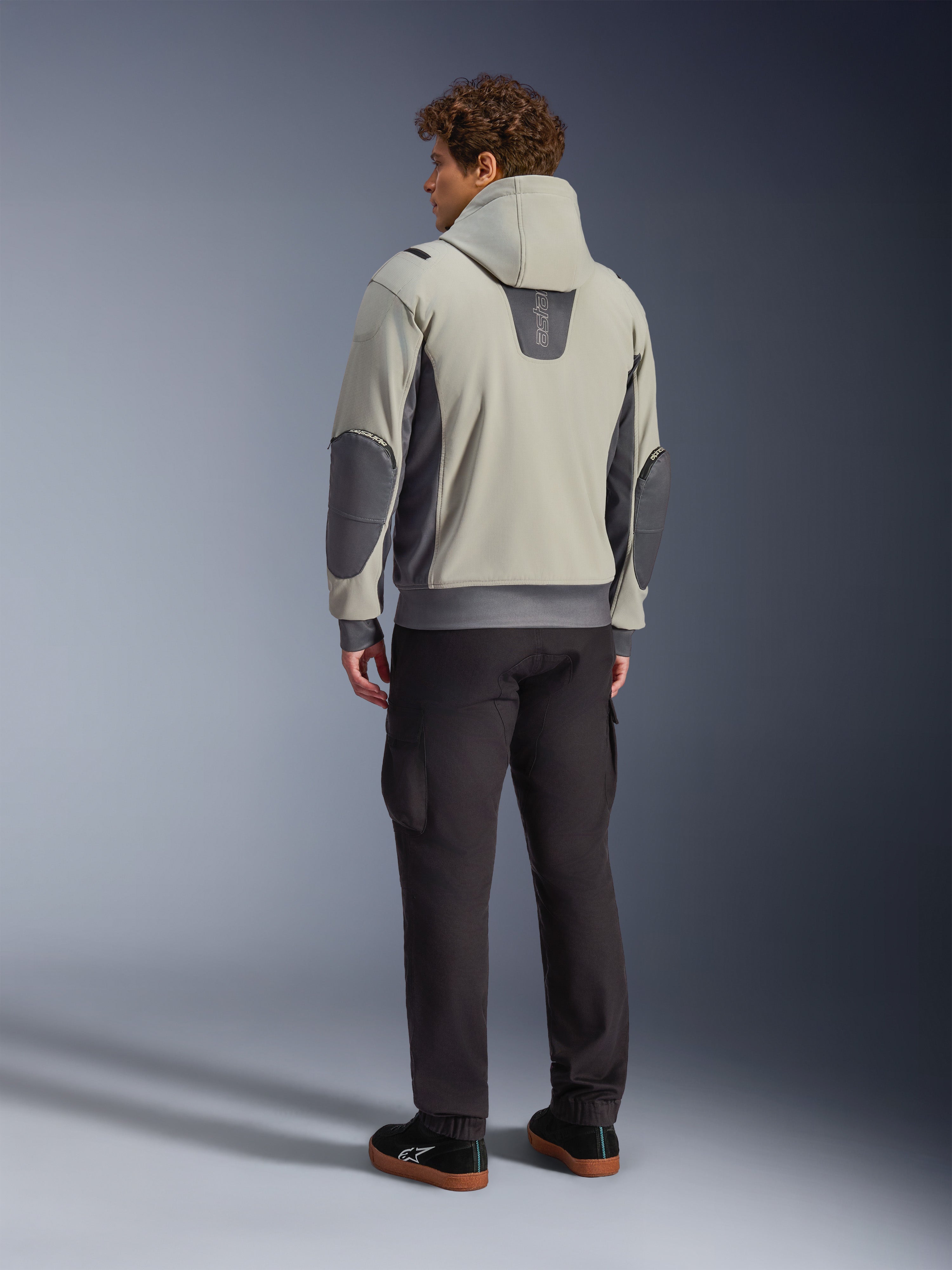 Modello visto di spalle che indossa la giacca Alpinestars Moblast Waterproof in Iron Gray Dark Sage, giacca da moto urbana con cappuccio con pannelli laterali grigio scuro e toppe rinforzate sui gomiti, abbinata a pantaloni da guida scuri e sneaker nere, estetica moderna e minimalista per il commuting urbano.