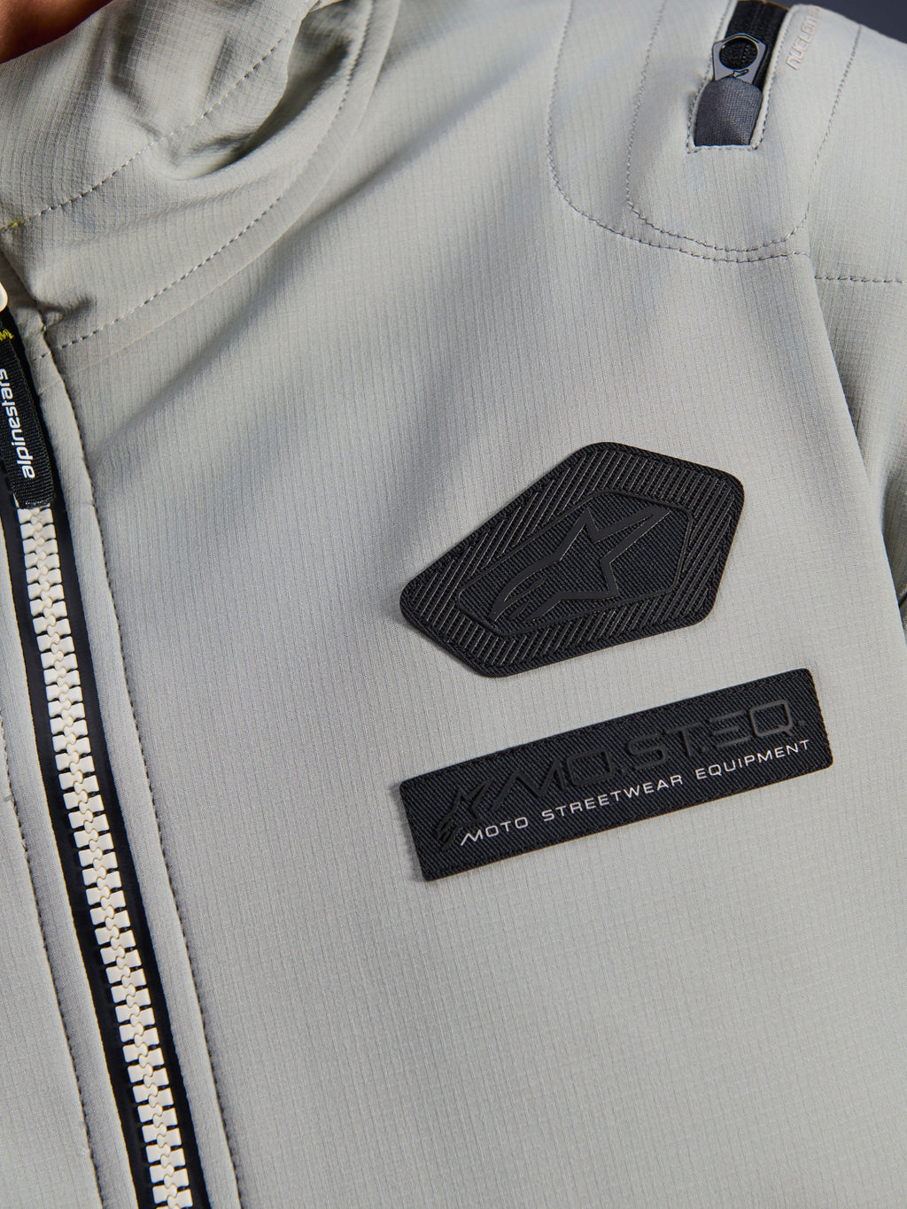 Giacca impermeabile Alpinestars Moblast, Iron Gray, dettaglio ravvicinato delle patch sul petto con logo Alpinestars e marchio MO.ST.EQ. Moto Streetwear Equipment, tessuto tecnico ripstop con robusta cerniera frontale e dettaglio della tasca sulla spalla.