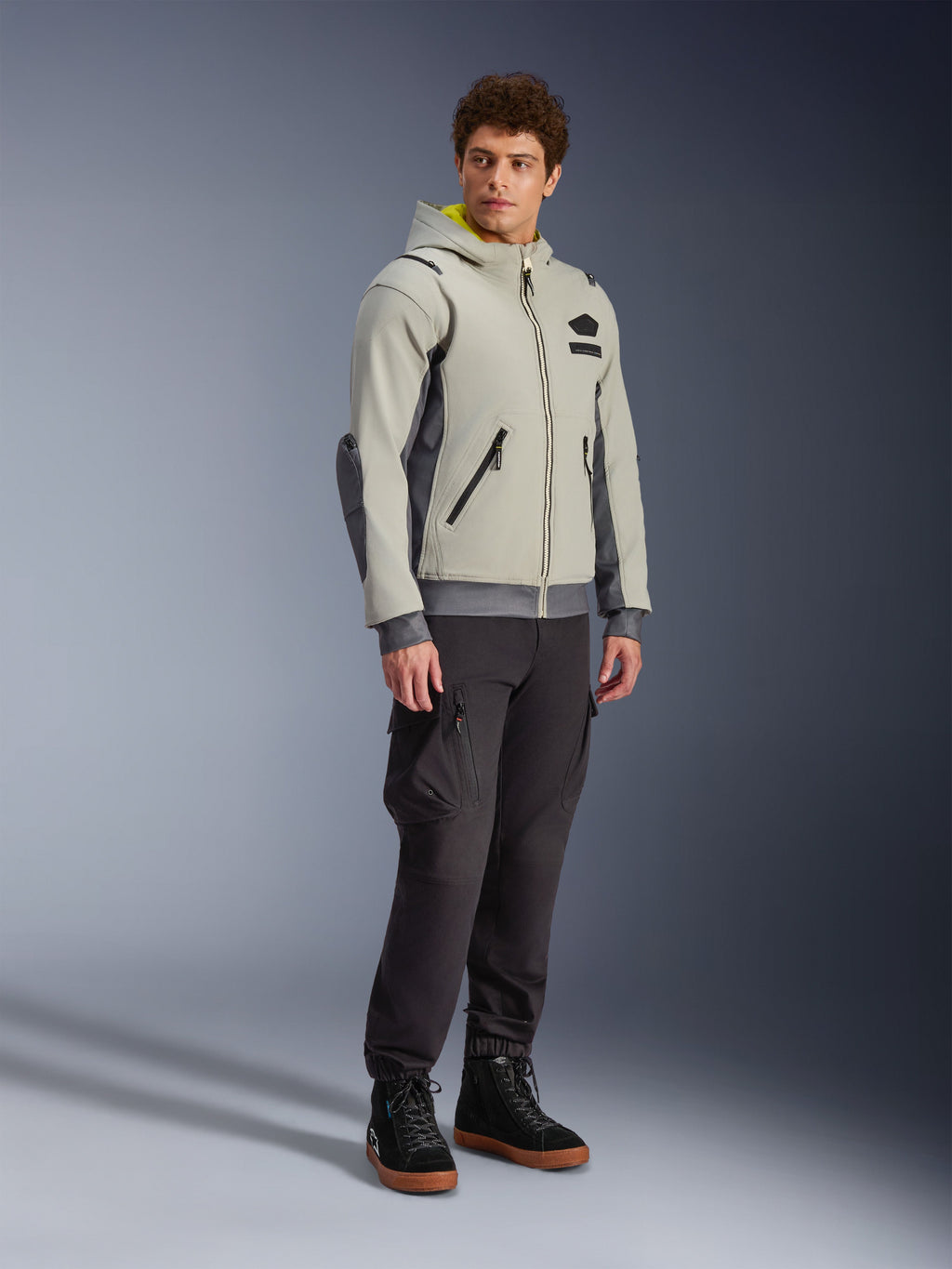 Alpinestars Moblast Waterproof Jacket, Iron Gray e Dark Sage, giacca da moto urbana con design a felpa con cappuccio, indossata da un modello con pantaloni da moto scuri e stivali neri, estetica cyber-tech minimalista con interno del cappuccio giallo e cerniere impermeabili