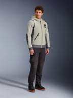 Alpinestars Moblast Waterproof Jacket, Iron Gray e Dark Sage, giacca da moto urbana con design a felpa con cappuccio, indossata da un modello con pantaloni da moto scuri e stivali neri, estetica cyber-tech minimalista con interno del cappuccio giallo e cerniere impermeabili