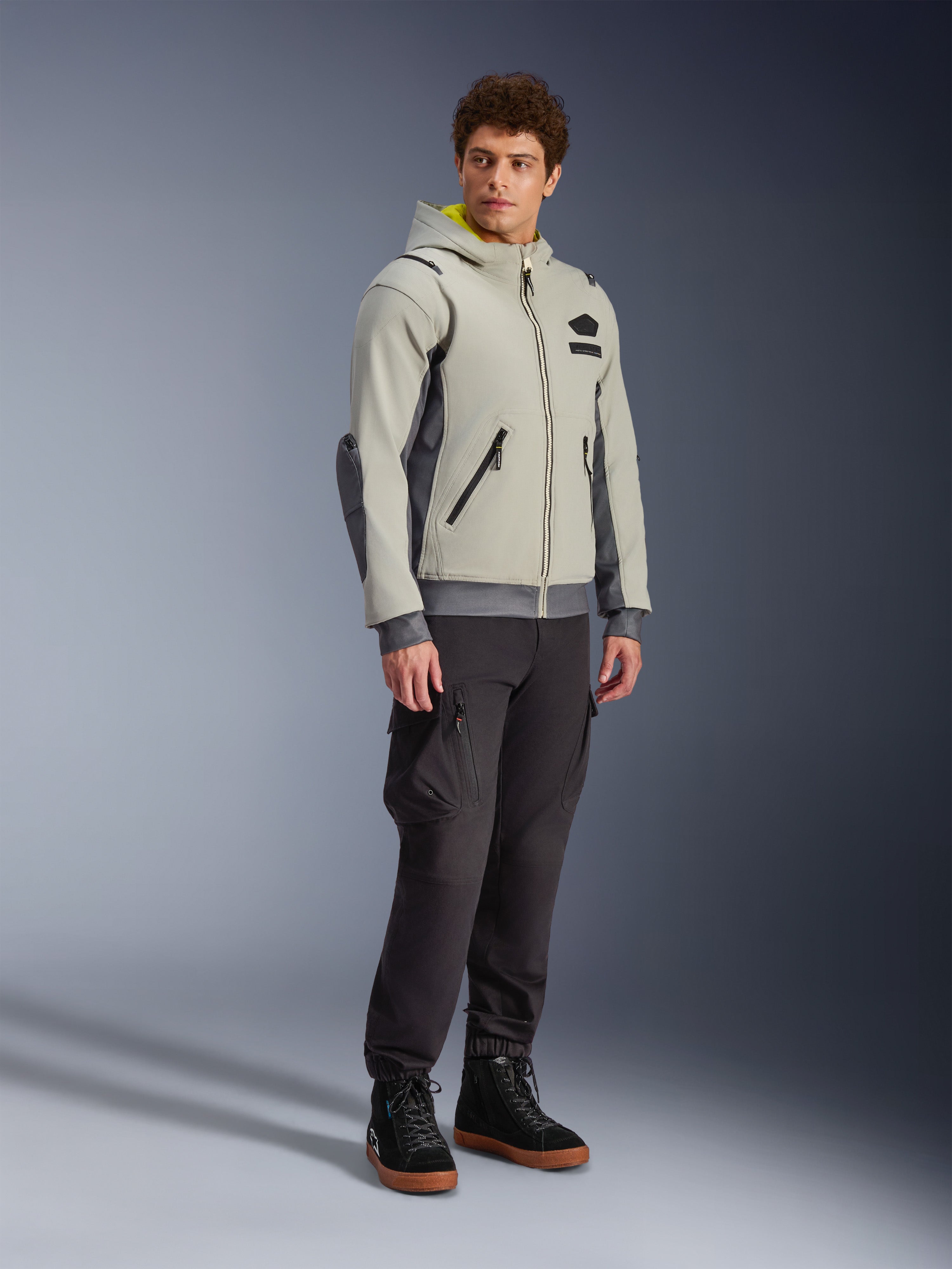 Alpinestars Moblast Waterproof Jacket, Iron Gray e Dark Sage, giacca da moto urbana con design a felpa con cappuccio, indossata da un modello con pantaloni da moto scuri e stivali neri, estetica cyber-tech minimalista con interno del cappuccio giallo e cerniere impermeabili