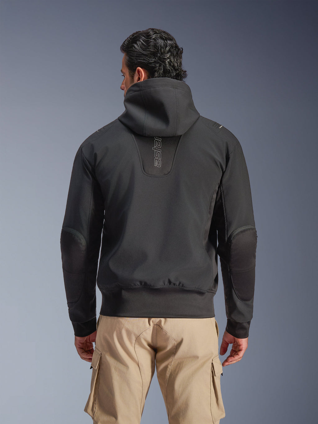 Alpinestars Giacca Moblast Waterproof, Nero, vista posteriore di giacca da moto urbana con cappuccio, con logo 'astars' verticale sulla schiena, vita e polsini elasticizzati, gomiti rinforzati e design moderno e tecnologico, indossata con pantaloni cargo beige