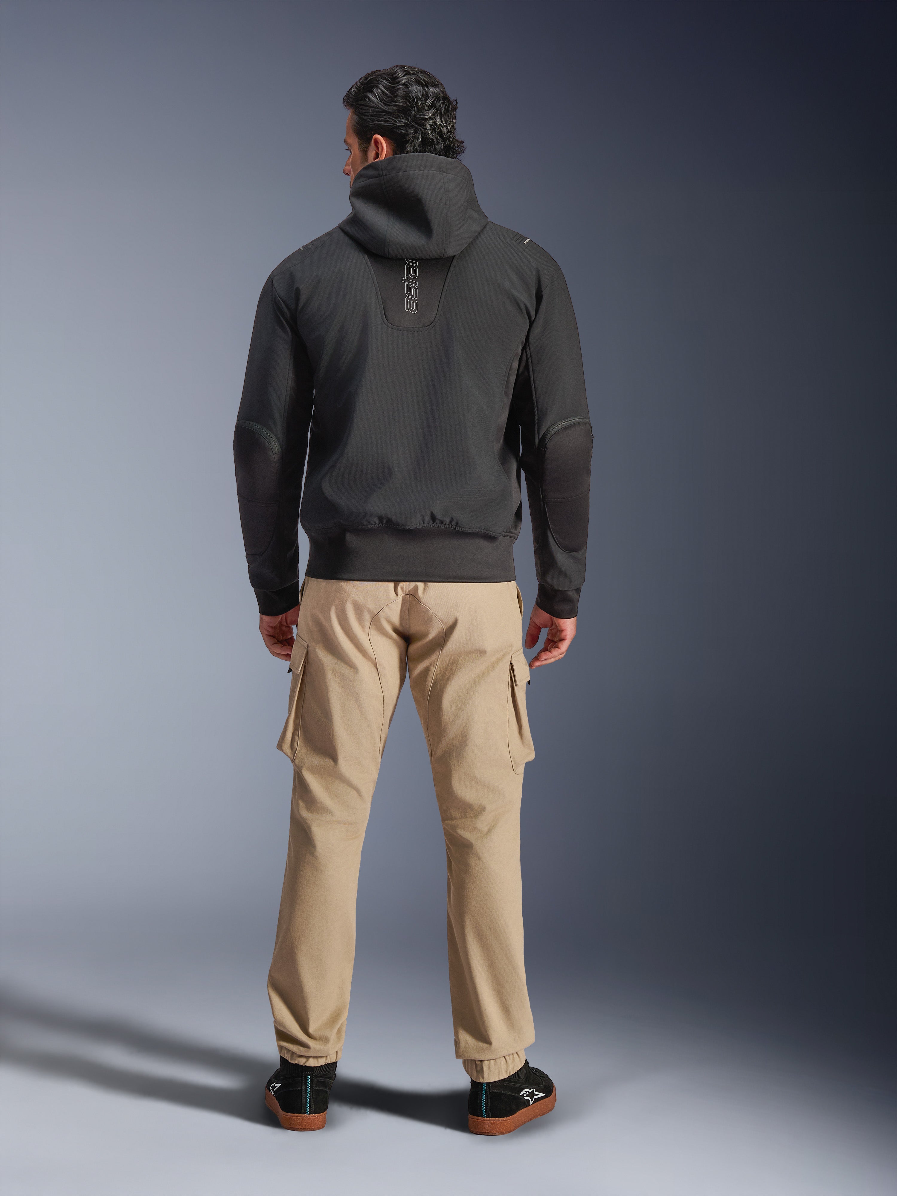 Alpinestars Moblast Waterproof Jacket, nero, vista posteriore di felpa da moto urbana con cappuccio, indossata con pantaloni cargo color sabbia e sneakers nere, design minimalista con marchio 'astars' verticale sulla parte superiore della schiena, abbigliamento tecnico da moto per ogni condizione atmosferica