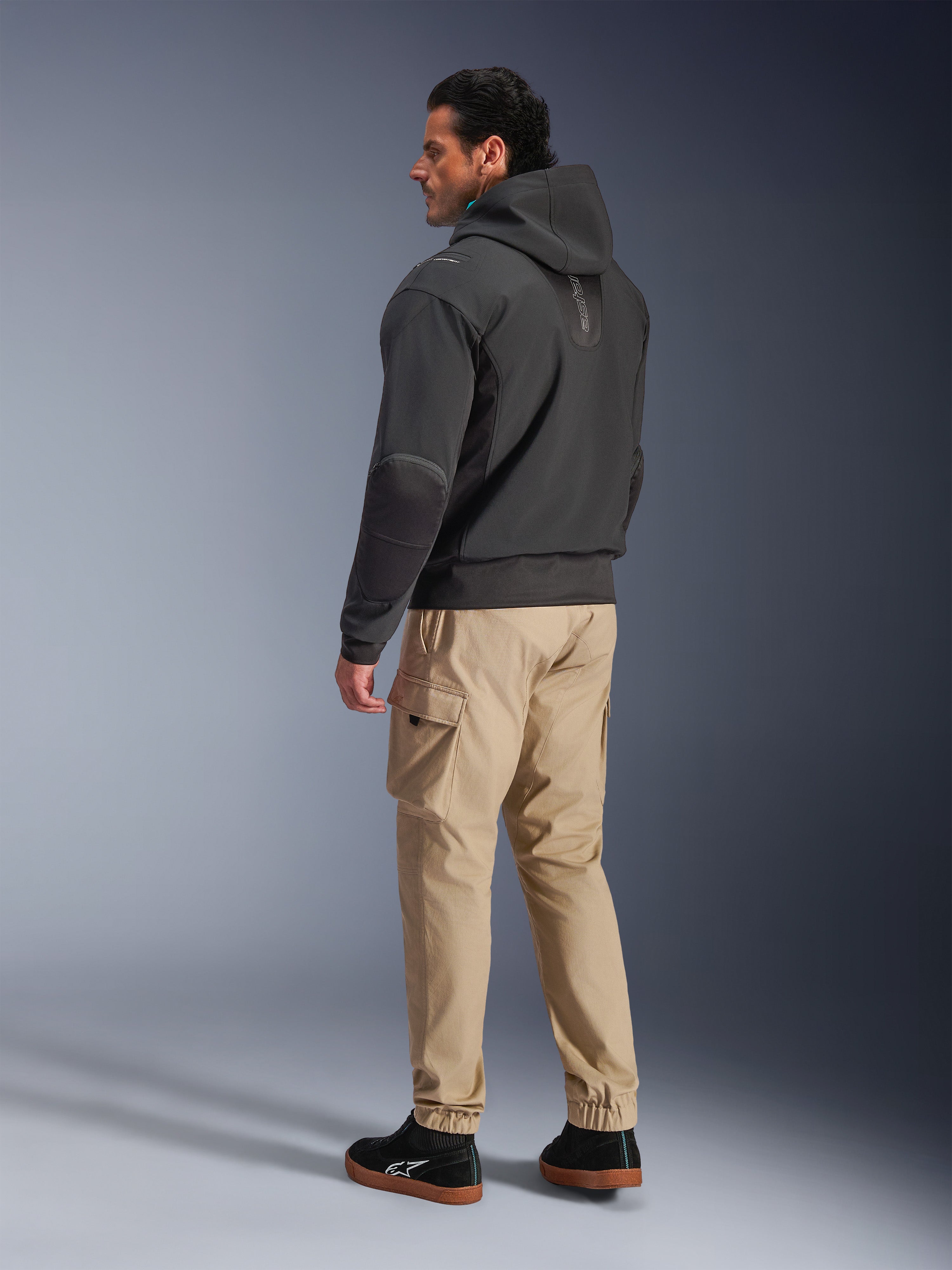 Giacca Alpinestars Moblast Waterproof, felpa tecnica da moto nera, vista laterale e posteriore, indossata con pantaloni cargo color tan e sneakers nere, con design con cappuccio, costruzione in tessuto tecnico ed estetica urbana moderna