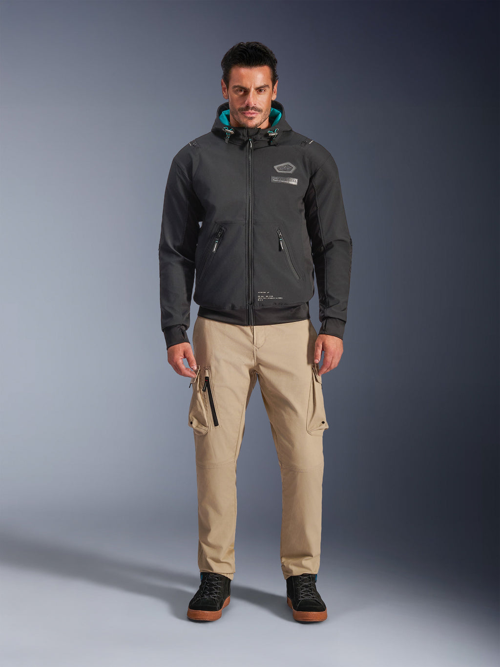 Giacca Alpinestars Moblast Waterproof, felpa da moto urbana nera con interno del cappuccio verde acqua, indossata con pantaloni cargo beige e stivali alti neri, abbigliamento da moto contemporaneo per tutte le stagioni