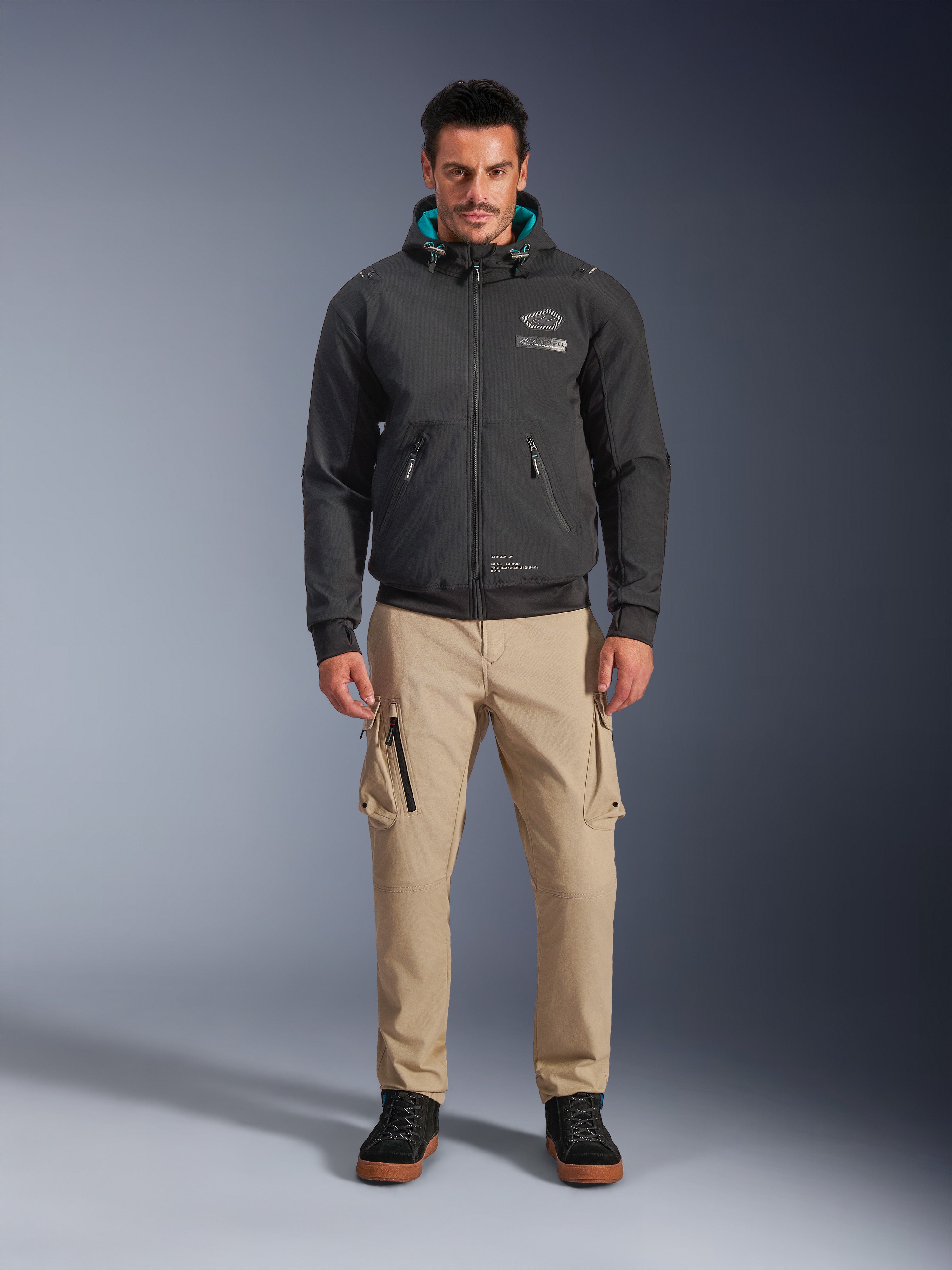 Giacca Alpinestars Moblast Waterproof, felpa da moto urbana nera con interno del cappuccio verde acqua, indossata con pantaloni cargo beige e stivali alti neri, abbigliamento da moto contemporaneo per tutte le stagioni