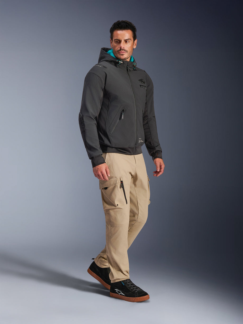 Modello che indossa la Giacca Alpinestars Moblast Waterproof in nero, felpa da moto urbana con cappuccio con fodera color ottanio, abbinata a pantaloni cargo kaki e sneakers nere, design moderno e minimalista in tessuto tecnico elasticizzato per il commuting in ogni condizione atmosferica