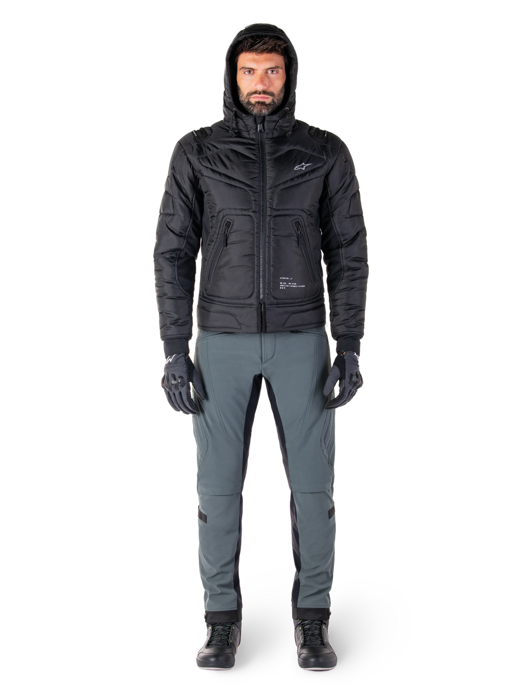 Giacca impermeabile Alpinestars Mohobbs in nero, modello che indossa un bomber trapuntato e imbottito con cappuccio, abbinato a pantaloni da moto grigi, guanti e stivali neri, abbigliamento da moto urban contemporary