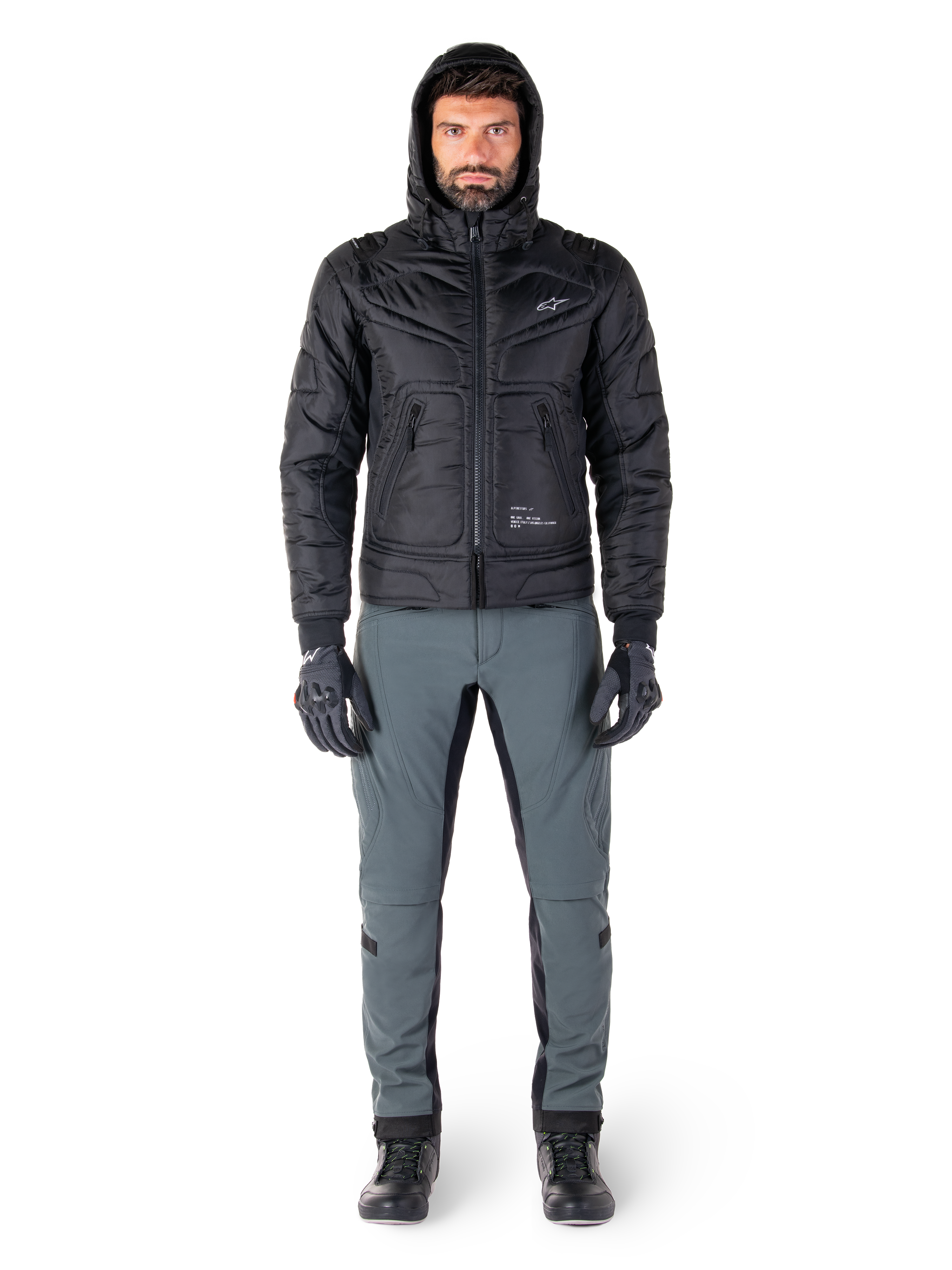 Giacca impermeabile Alpinestars Mohobbs in nero, modello che indossa un bomber trapuntato e imbottito con cappuccio, abbinato a pantaloni da moto grigi, guanti e stivali neri, abbigliamento da moto urban contemporary