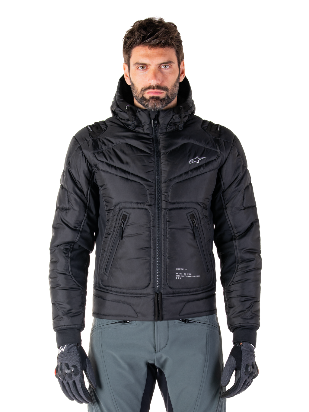 Alpinestars Mohobbs Waterproof Jacket, giacca da moto bomber imbottita nera, tessuto in poliestere trapuntato con cerniera frontale e tasche con zip, piccolo logo bianco sul petto, indossata da un modello con pantaloni grigi e guanti, design urban streetwear per la guida in città.