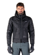 Alpinestars Mohobbs Waterproof Jacket, giacca da moto bomber imbottita nera, tessuto in poliestere trapuntato con cerniera frontale e tasche con zip, piccolo logo bianco sul petto, indossata da un modello con pantaloni grigi e guanti, design urban streetwear per la guida in città.