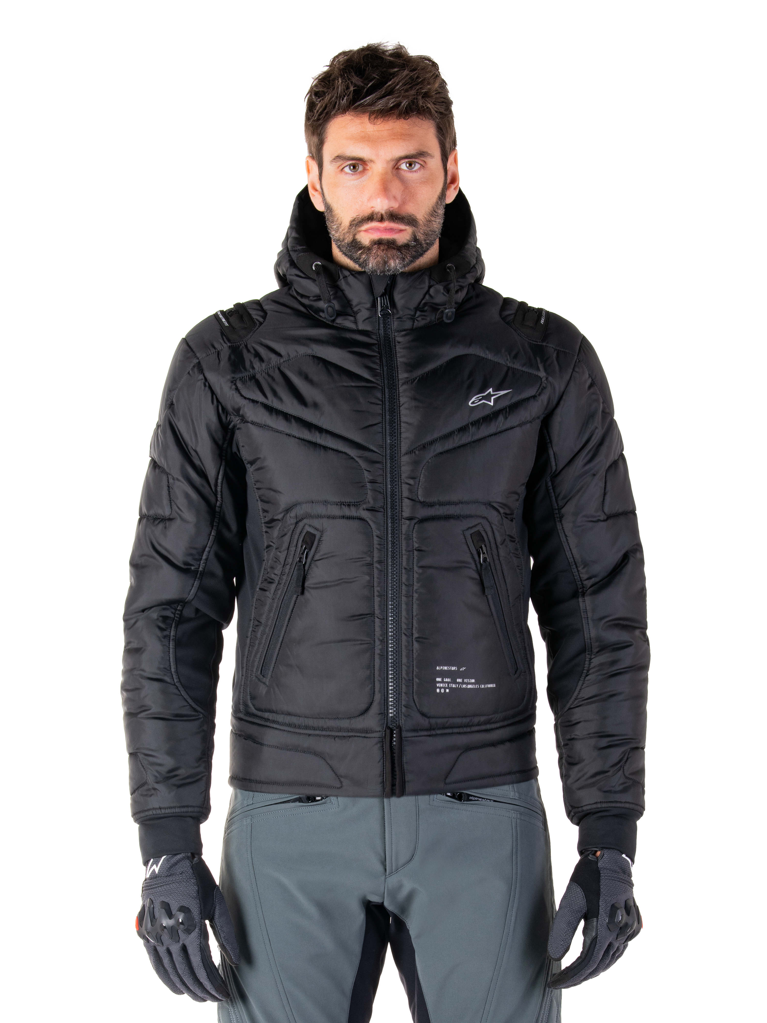 Alpinestars Mohobbs Waterproof Jacket, giacca da moto bomber imbottita nera, tessuto in poliestere trapuntato con cerniera frontale e tasche con zip, piccolo logo bianco sul petto, indossata da un modello con pantaloni grigi e guanti, design urban streetwear per la guida in città.