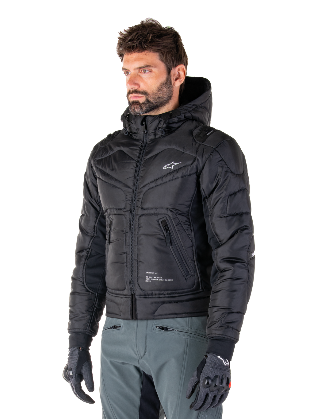 Giacca impermeabile Alpinestars Mohobbs, nero, design bomber trapuntato e imbottito con cappuccio, indossata da un uomo con pantaloni da moto grigi e guanti neri, abbigliamento da moto ispirato allo streetwear urbano con un piccolo logo bianco sul petto.