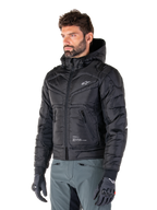 Giacca impermeabile Alpinestars Mohobbs, nero, design bomber trapuntato e imbottito con cappuccio, indossata da un uomo con pantaloni da moto grigi e guanti neri, abbigliamento da moto ispirato allo streetwear urbano con un piccolo logo bianco sul petto.