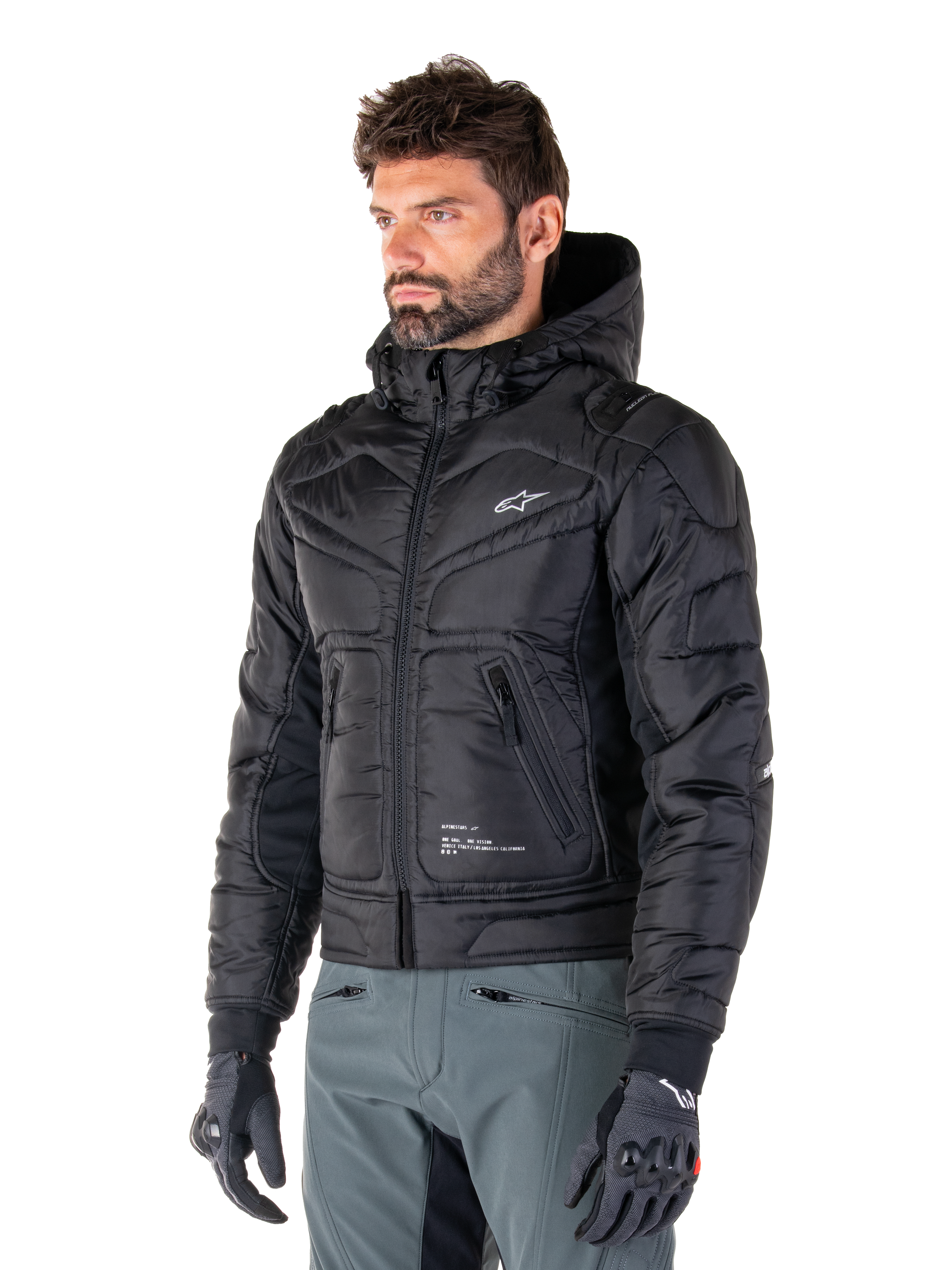 Giacca impermeabile Alpinestars Mohobbs, nero, design bomber trapuntato e imbottito con cappuccio, indossata da un uomo con pantaloni da moto grigi e guanti neri, abbigliamento da moto ispirato allo streetwear urbano con un piccolo logo bianco sul petto.