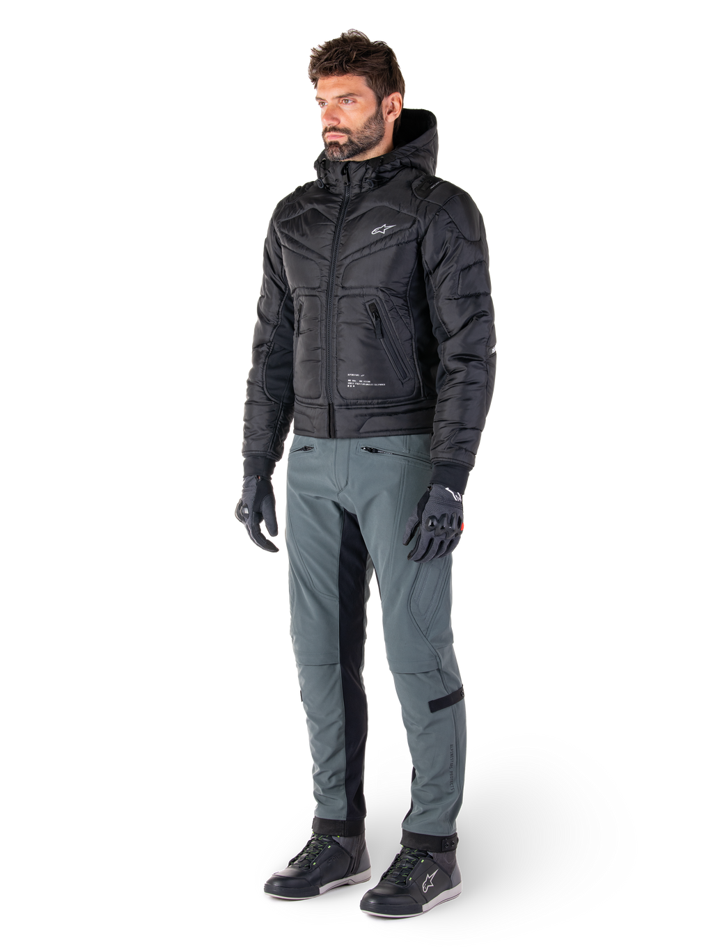 Alpinestars Mohobbs Waterproof Jacket, bomber da moto urban trapuntato nero con cappuccio, indossata da un modello con pantaloni da guida grigi e guanti neri, con logo bianco sul petto, abbigliamento protettivo contemporaneo d'ispirazione streetwear