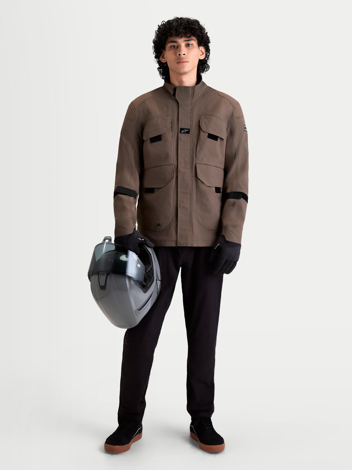 Alpinestars Altan Drystar Giacca, giacca da moto urbana contemporanea da uomo in marrone noce, vista frontale su un modello che indossa pantaloni neri, guanti e tiene in mano un casco, struttura in tessuto impermeabile con tasche cargo