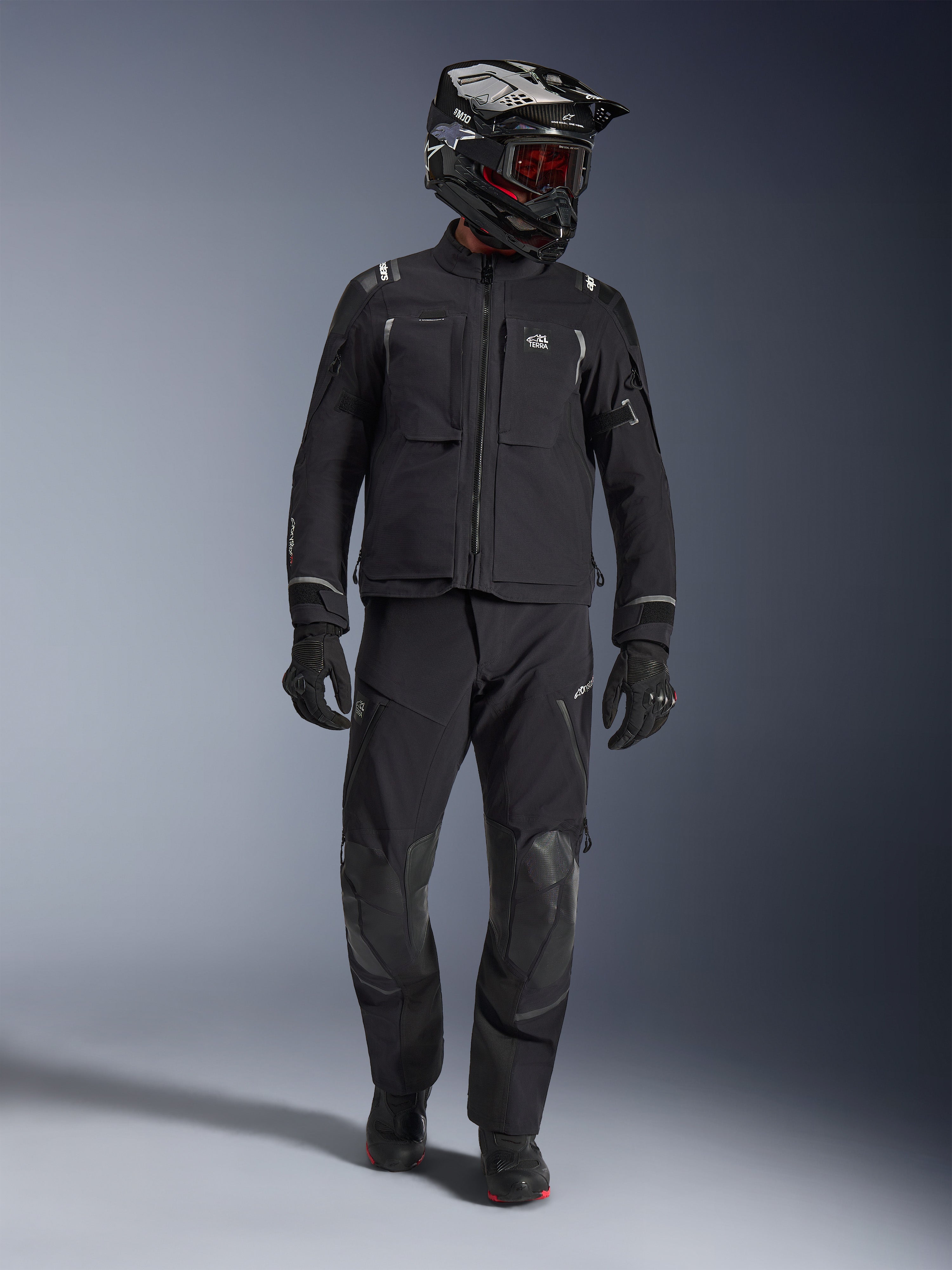 Pilota adventure che indossa la giacca Alpinestars Andes Pro Drystar® XF in nero e grigio scuro, pantaloni adventure neri coordinati, casco bianco e nero con maschera rossa, guanti e stivali neri; presenta ampie tasche anteriori, dettagli riflettenti e costruzione tecnica laminata a 3 strati per il turismo in ogni condizione climatica.