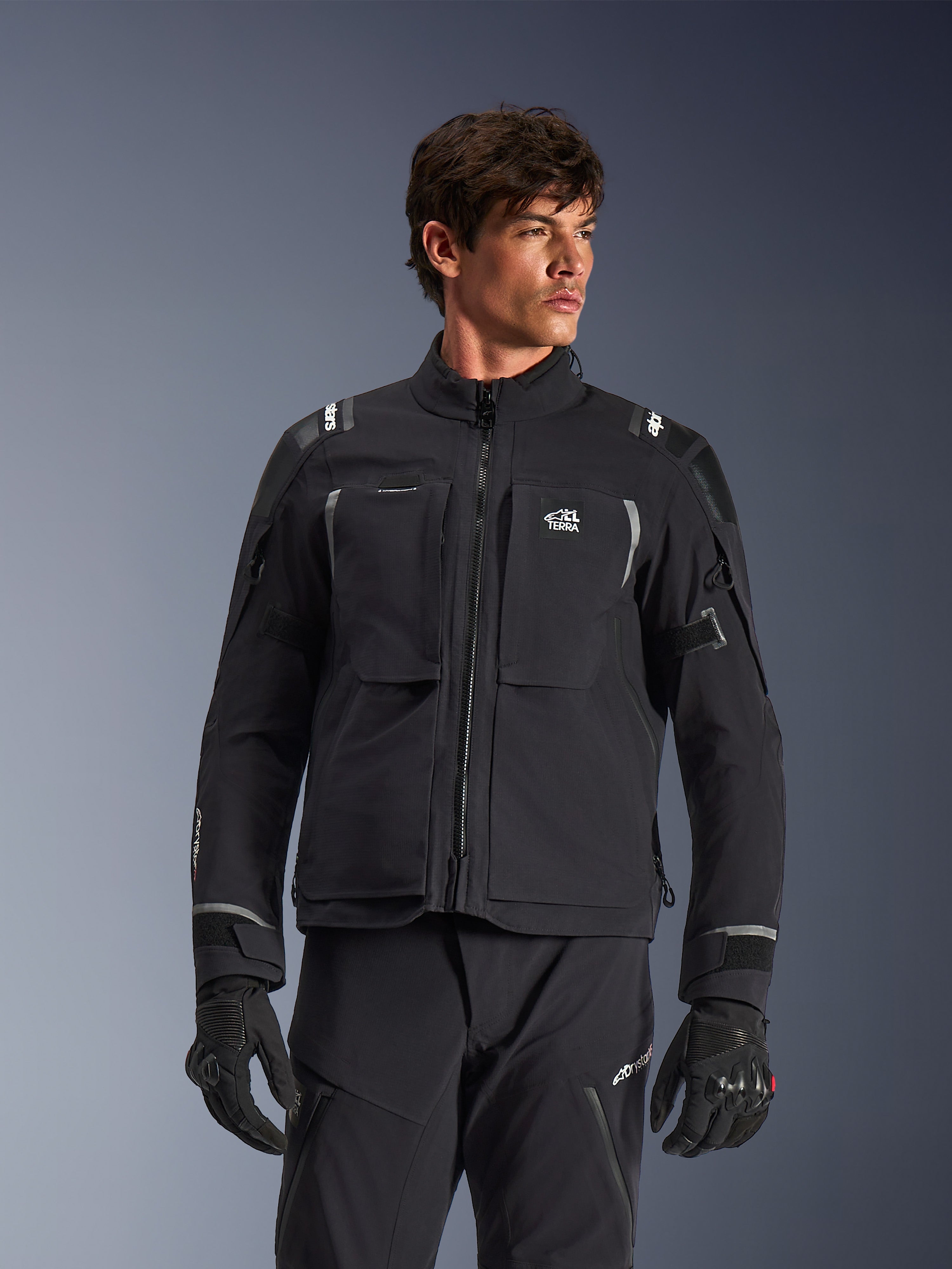 Giacca Alpinestars Andes Pro Drystar® XF, giacca da moto adventure touring nera e grigio scuro, indossata con pantaloni e guanti coordinati, caratterizzata da una struttura tecnica a guscio laminato, logo ALL TERRA sul petto, dettagli riflettenti e polsini regolabili
