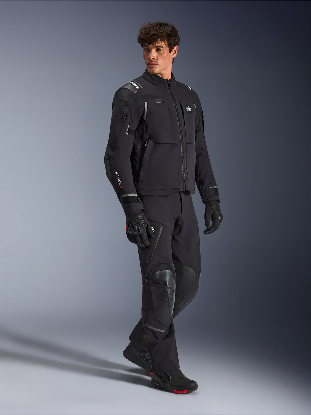 Giacca Alpinestars Andes Pro Drystar® XF, giacca da moto nera e grigio scuro, indossata da un modello con pantaloni adventure coordinati, guanti e stivali, scatto in studio a figura intera, abbigliamento tecnico da touring adventure con dettagli riflettenti e branding