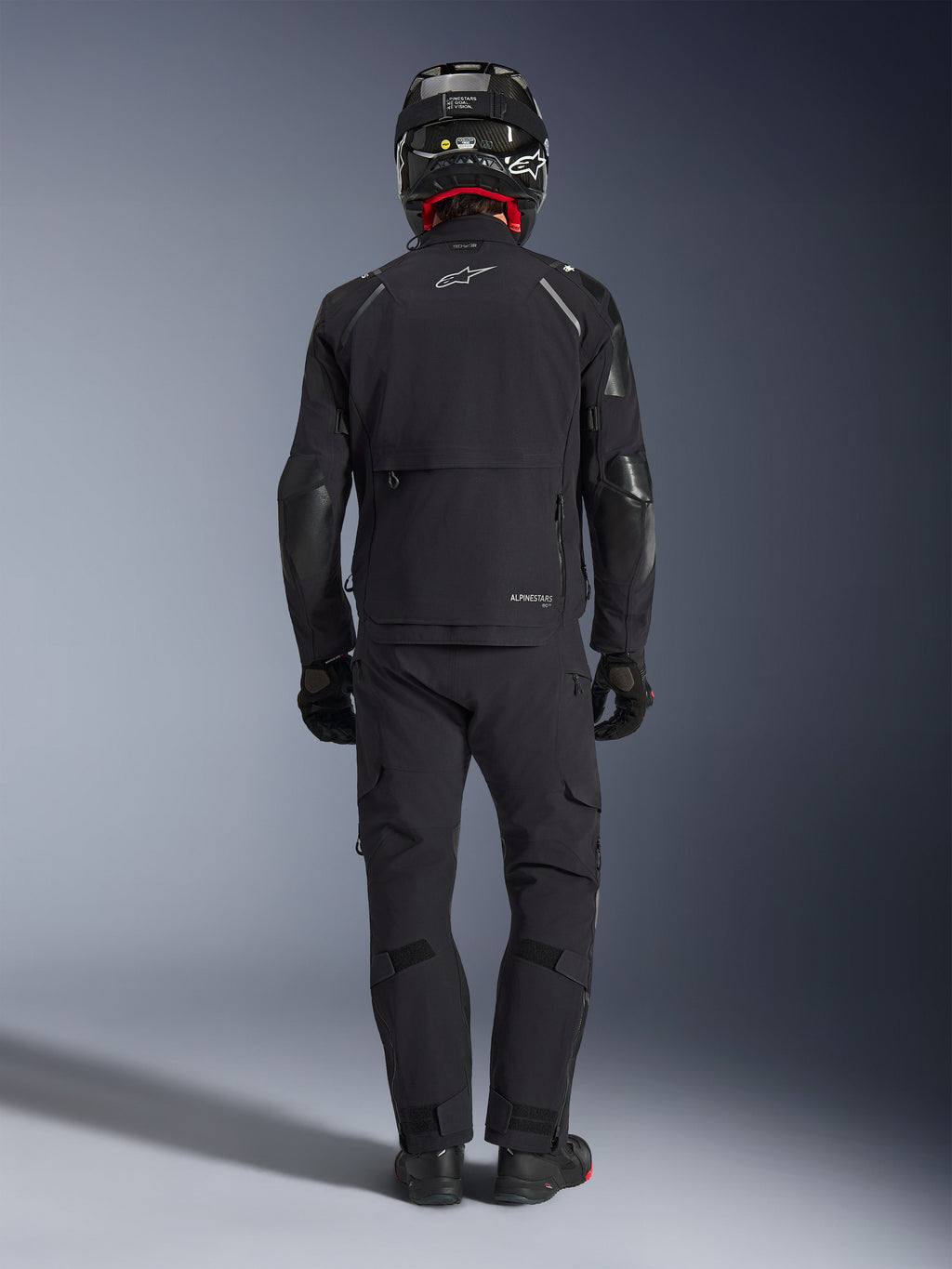 Giacca Alpinestars Andes Pro Drystar® XF in nero e grigio scuro, vista posteriore di un pilota con abbigliamento adventure touring completo, pantaloni abbinati e casco in fibra di carbonio, dotata di ampia tasca cargo posteriore e loghi Alpinestars riflettenti.