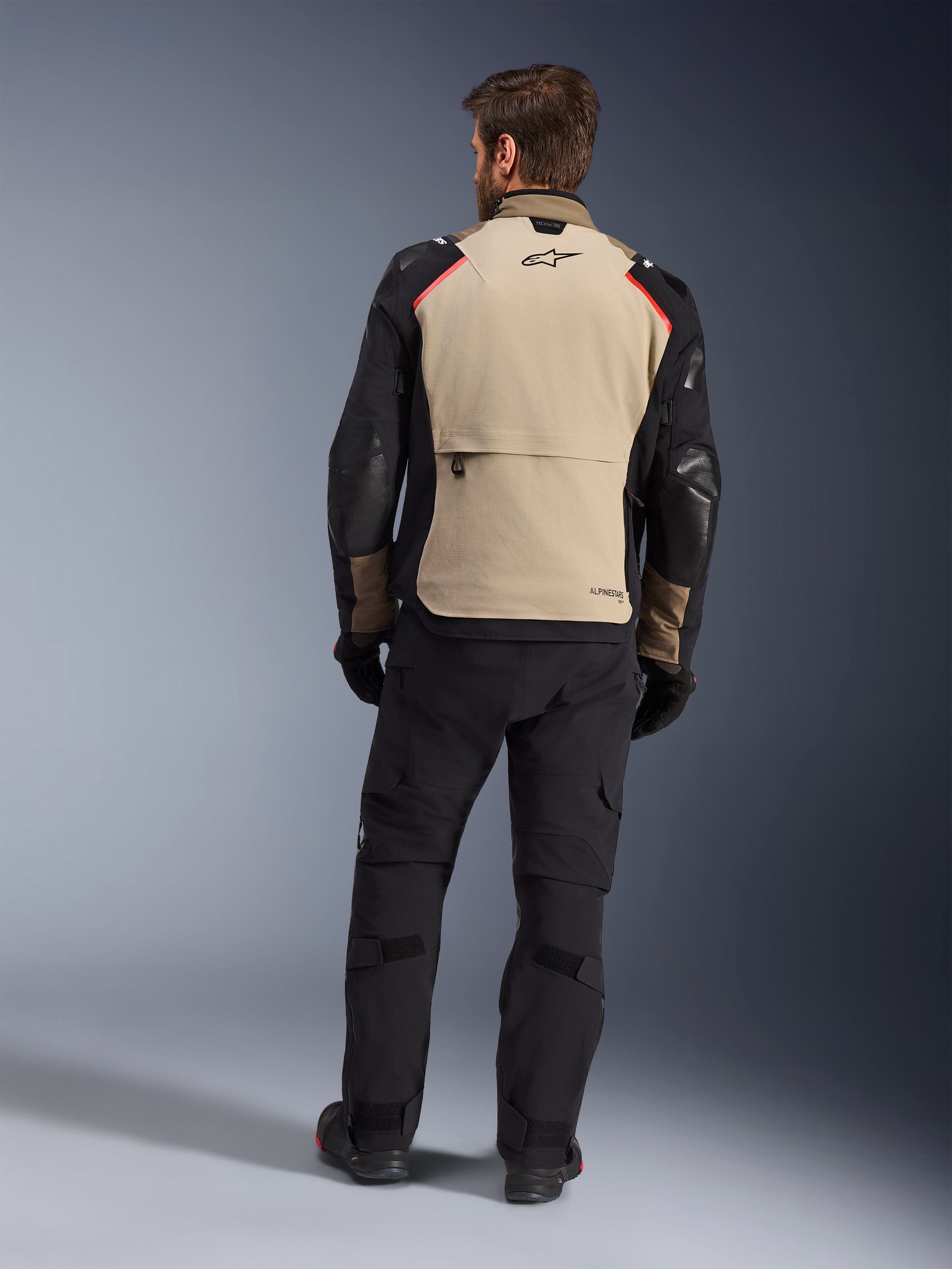 Giacca Alpinestars Andes Pro Drystar® XF, Walnut Aluminum, vista posteriore, giacca da moto adventure con maniche nere e dettagli rossi sulle spalle, indossata con pantaloni da moto e guanti neri
