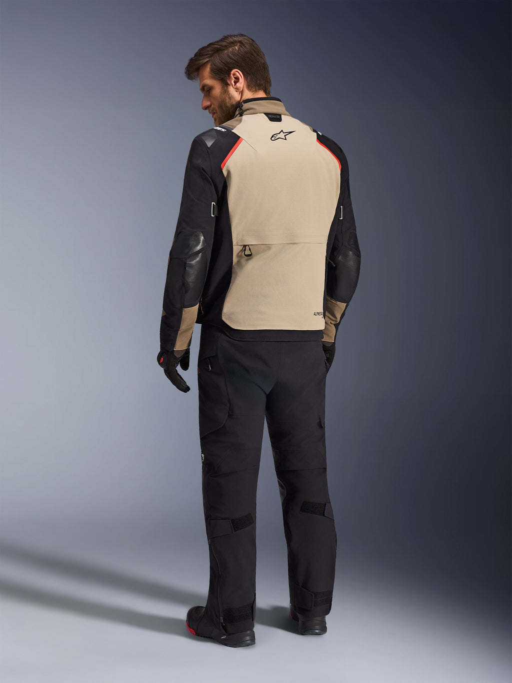 Alpinestars Andes Pro Drystar® XF Jacket, giacca da moto, color noce e nero con accenti rossi, vista posteriore su modello, design adventure touring con ampia tasca posteriore portaoggetti, indossata con pantaloni da moto e guanti neri.
