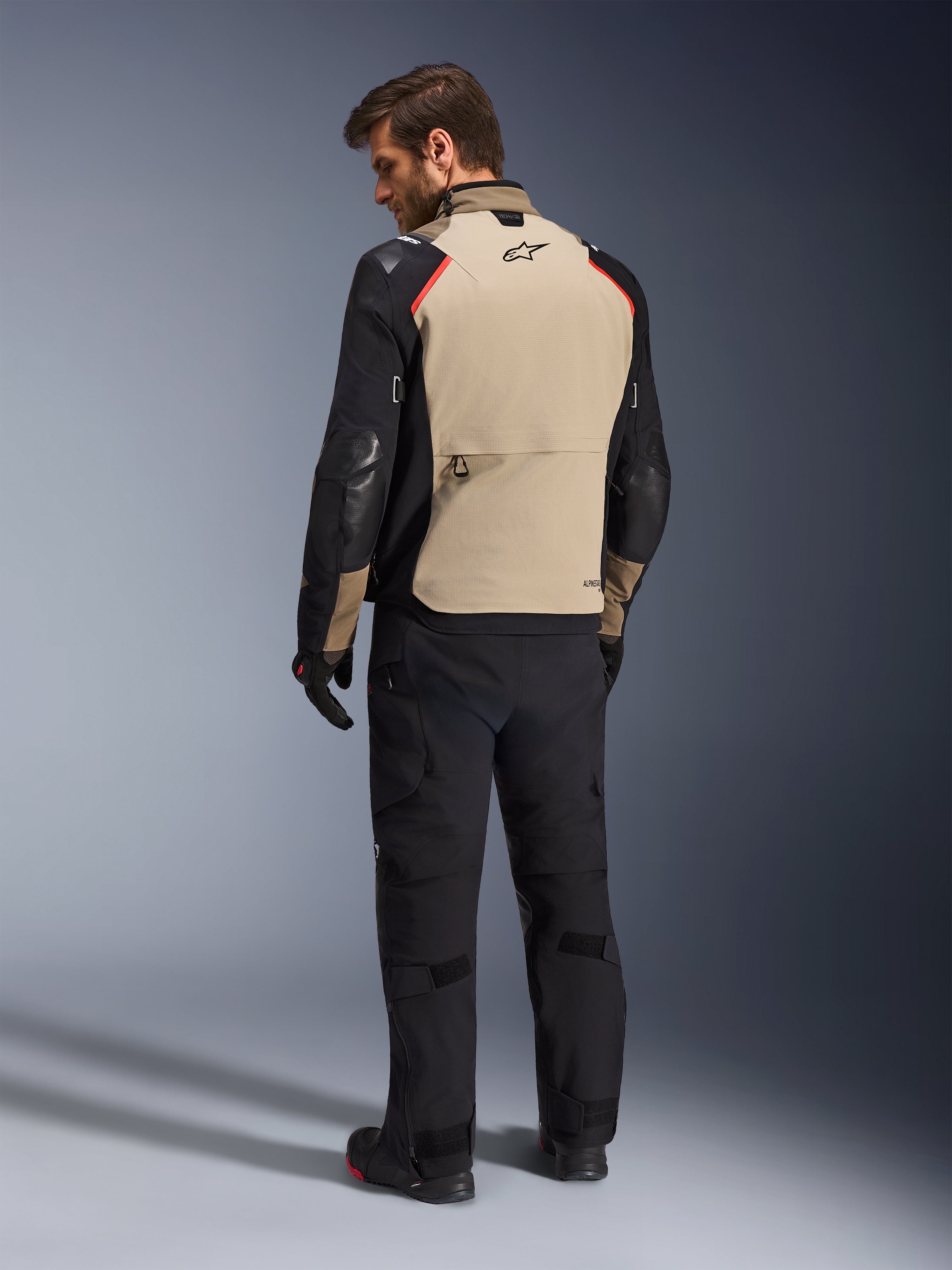 Alpinestars Andes Pro Drystar® XF Jacket, giacca da moto, color noce e nero con accenti rossi, vista posteriore su modello, design adventure touring con ampia tasca posteriore portaoggetti, indossata con pantaloni da moto e guanti neri.