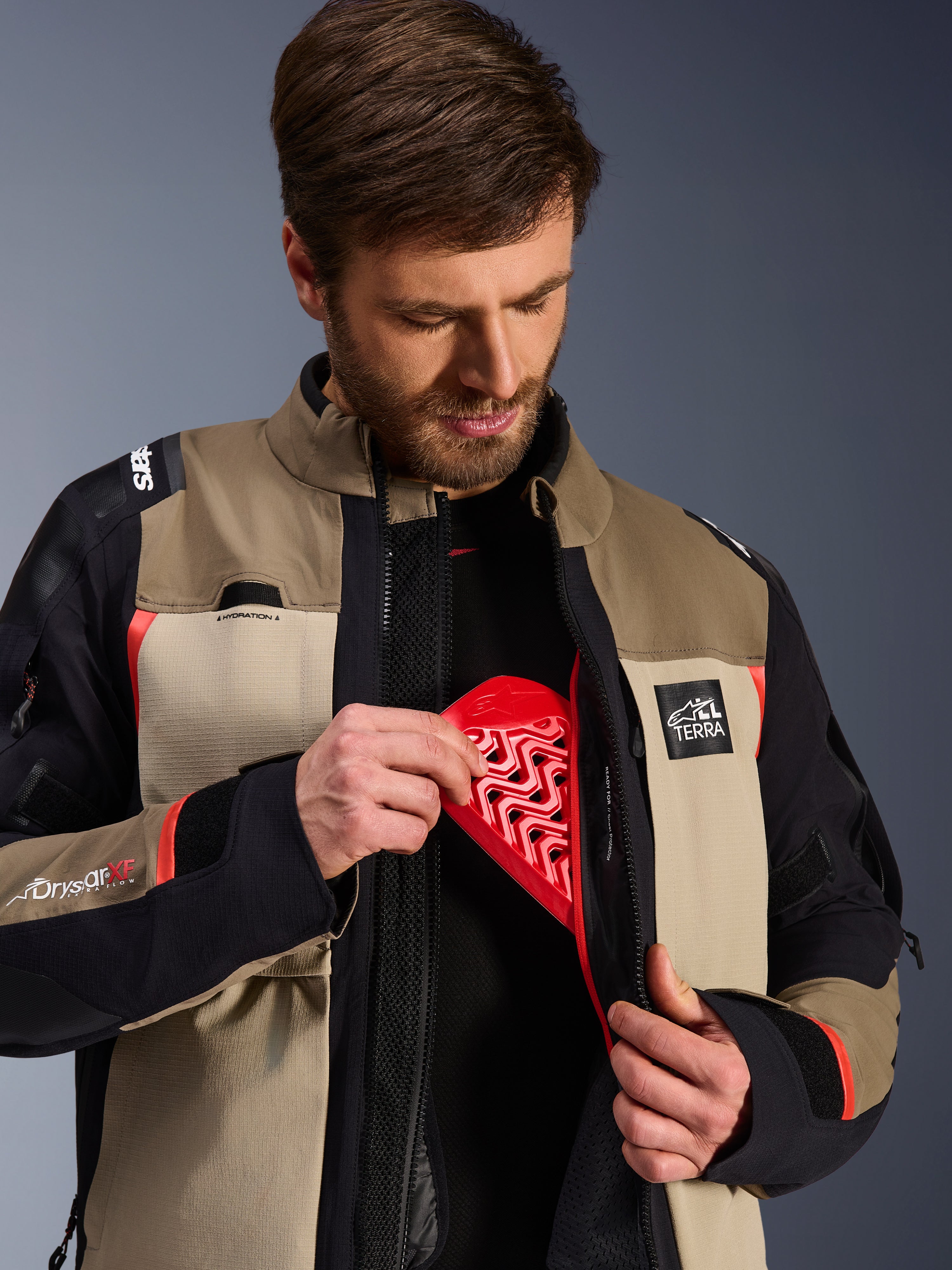 Giacca Alpinestars Andes Pro Drystar® XF in Walnut Aluminum, modello che mostra l'inserimento della protezione per il torace Nucleon rossa nella tasca interna, giacca da moto adventure con marchio ALL TERRA e loghi Drystar XF