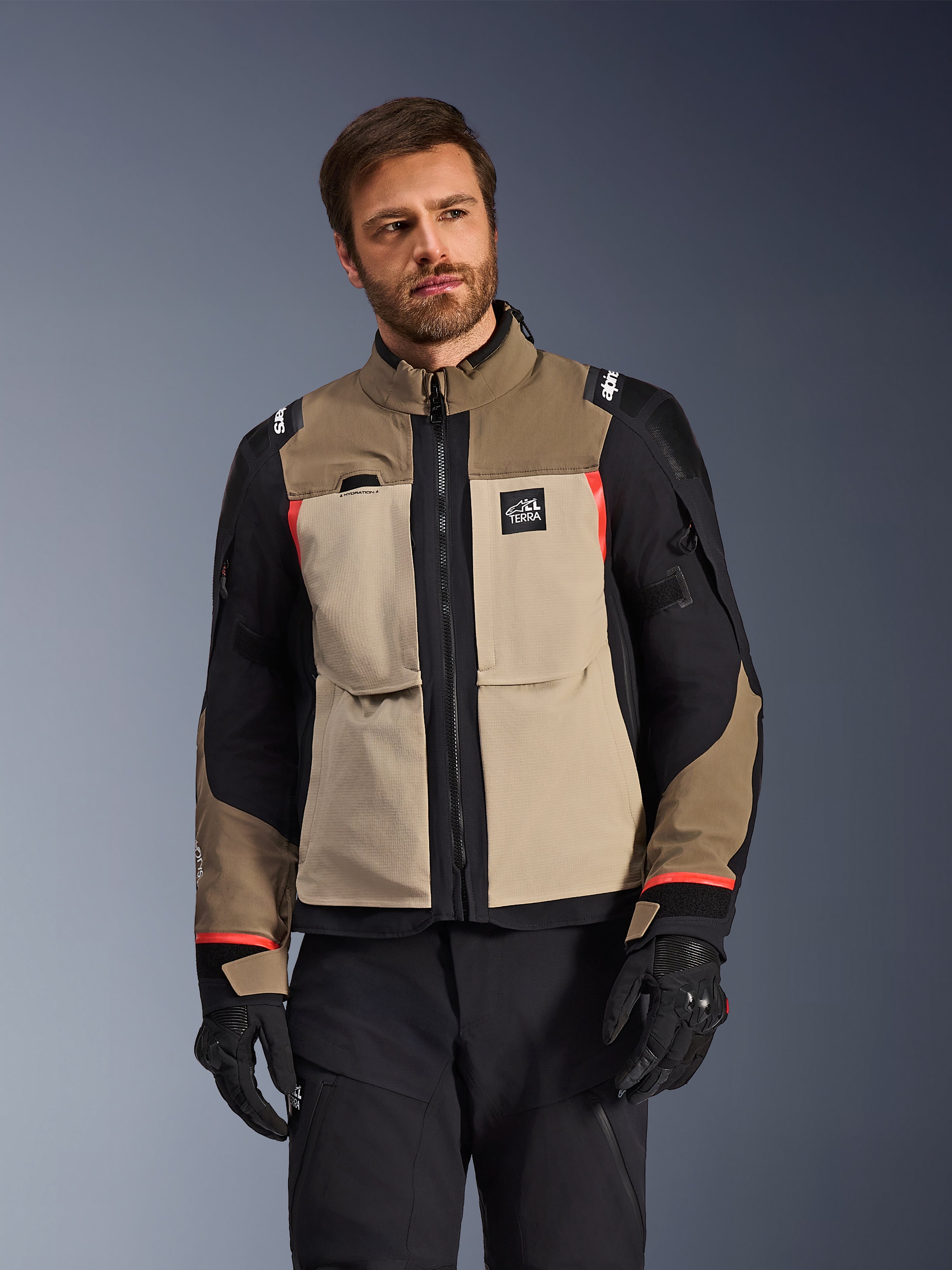 Giacca Alpinestars Andes Pro Drystar® XF, Walnut Aluminum, giacca da moto adventure con pannelli color sabbia e neri e accenti rossi, indossata con pantaloni da guida e guanti neri, abbigliamento tecnico da touring impermeabile