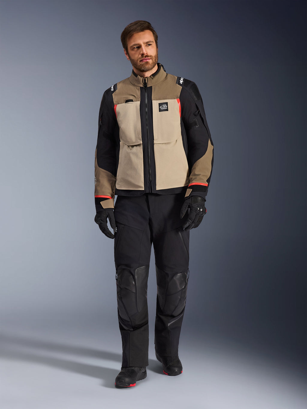 Modello con giacca Alpinestars Andes Pro Drystar® XF in Walnut Aluminum, giacca da touring adventure marrone e nera con accenti rossi, abbinata a pantaloni da moto, guanti e stivali neri, scatto in studio a figura intera per abbigliamento protettivo.