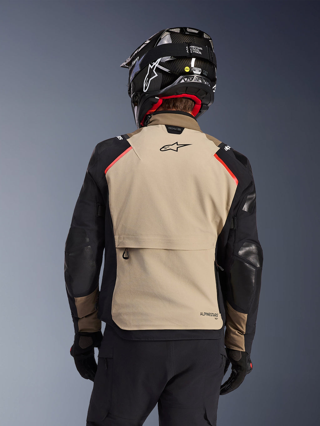 Giacca Alpinestars Andes Pro Drystar® XF in Walnut Aluminum, vista posteriore, pannello posteriore color sabbia con ampia tasca cargo, maniche nere con pannelli rinforzati, logo Alpinestars sulla parte superiore della schiena, abbinata a casco e guanti neri per l'adventure touring.