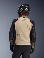 Giacca Alpinestars Andes Pro Drystar® XF in Walnut Aluminum, vista posteriore, pannello posteriore color sabbia con ampia tasca cargo, maniche nere con pannelli rinforzati, logo Alpinestars sulla parte superiore della schiena, abbinata a casco e guanti neri per l'adventure touring.