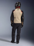 Pilota visto da dietro che indossa la giacca Alpinestars Andes Pro Drystar® XF in Walnut Aluminum, pannello posteriore color sabbia con maniche nere e dettagli rossi, abbinata a pantaloni tecnici adventure neri e casco da moto, equipaggiamento touring premium per tutte le stagioni