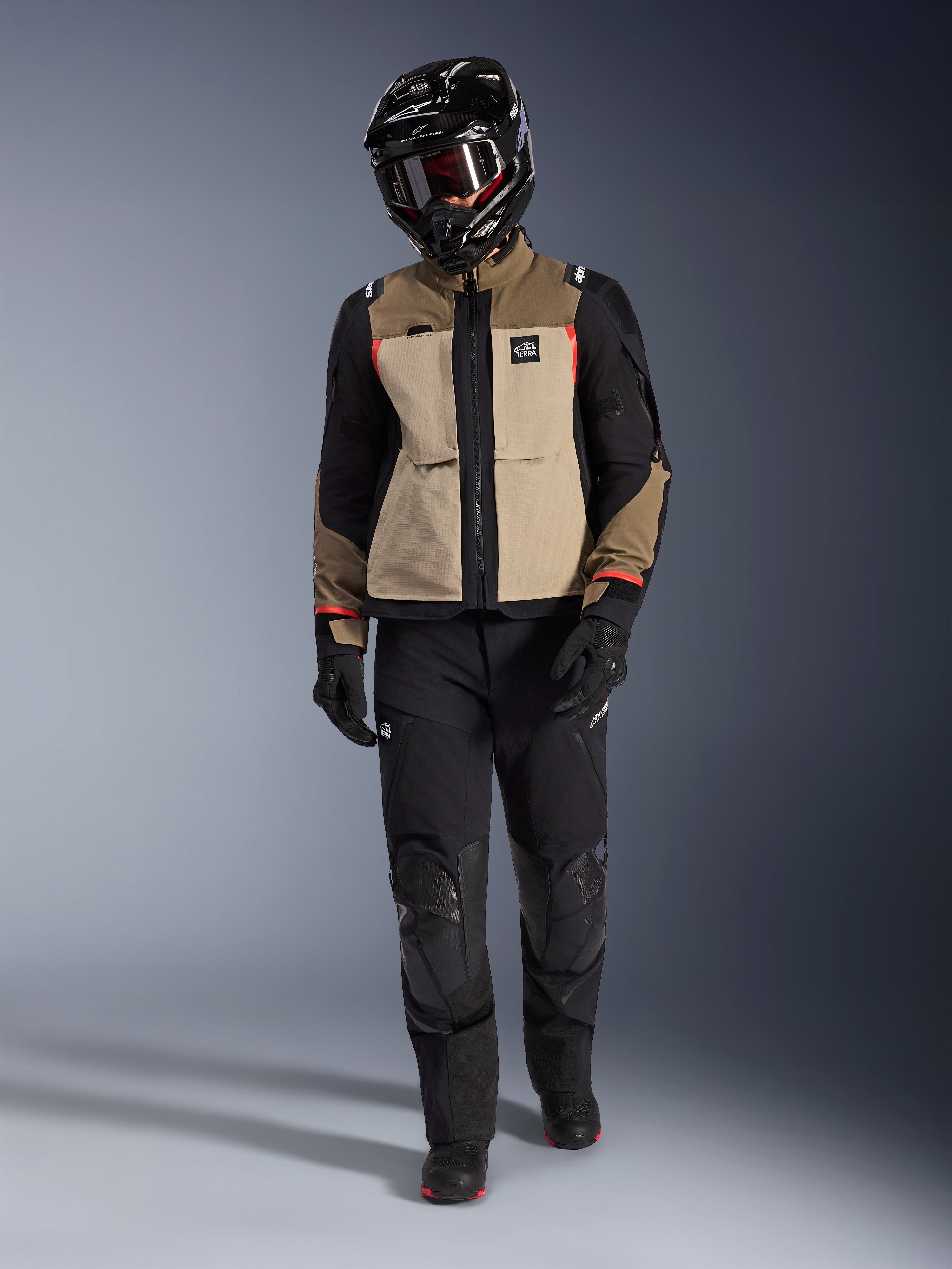 Giacca Alpinestars Andes Pro Drystar® XF in Walnut Aluminum, indossata con pantaloni adventure neri, guanti neri e casco integrale nero con maschera, con dettagli rossi e marchio All Terra, set-up professionale per adventure touring