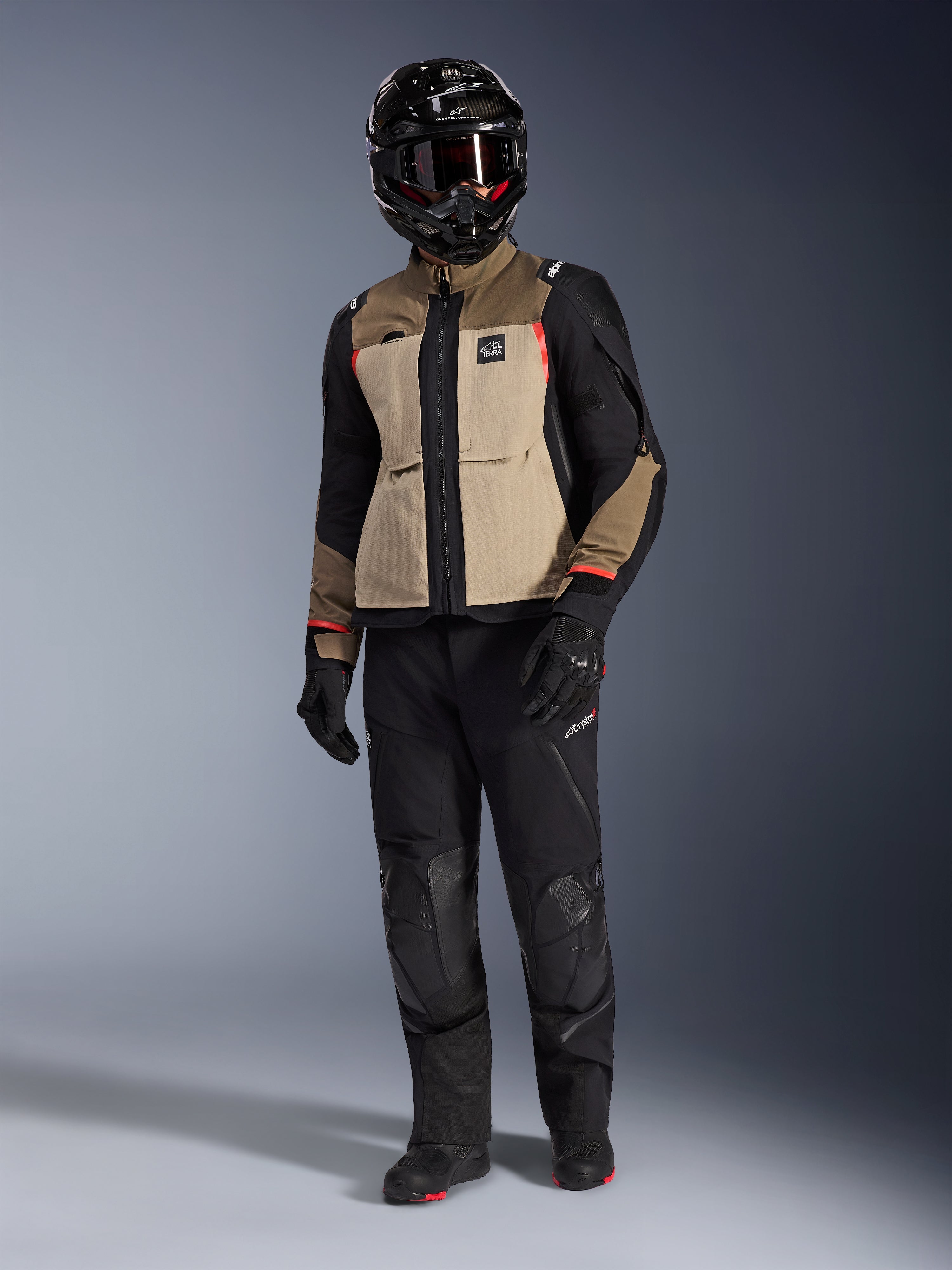 Alpinestars Andes Pro Drystar® XF Jacket in Walnut Aluminum, giacca da moto adventure indossata con pantaloni neri abbinati, casco adventure nero con maschera e stivali neri, caratterizzata da pannelli color sabbia e neri con dettagli rossi.