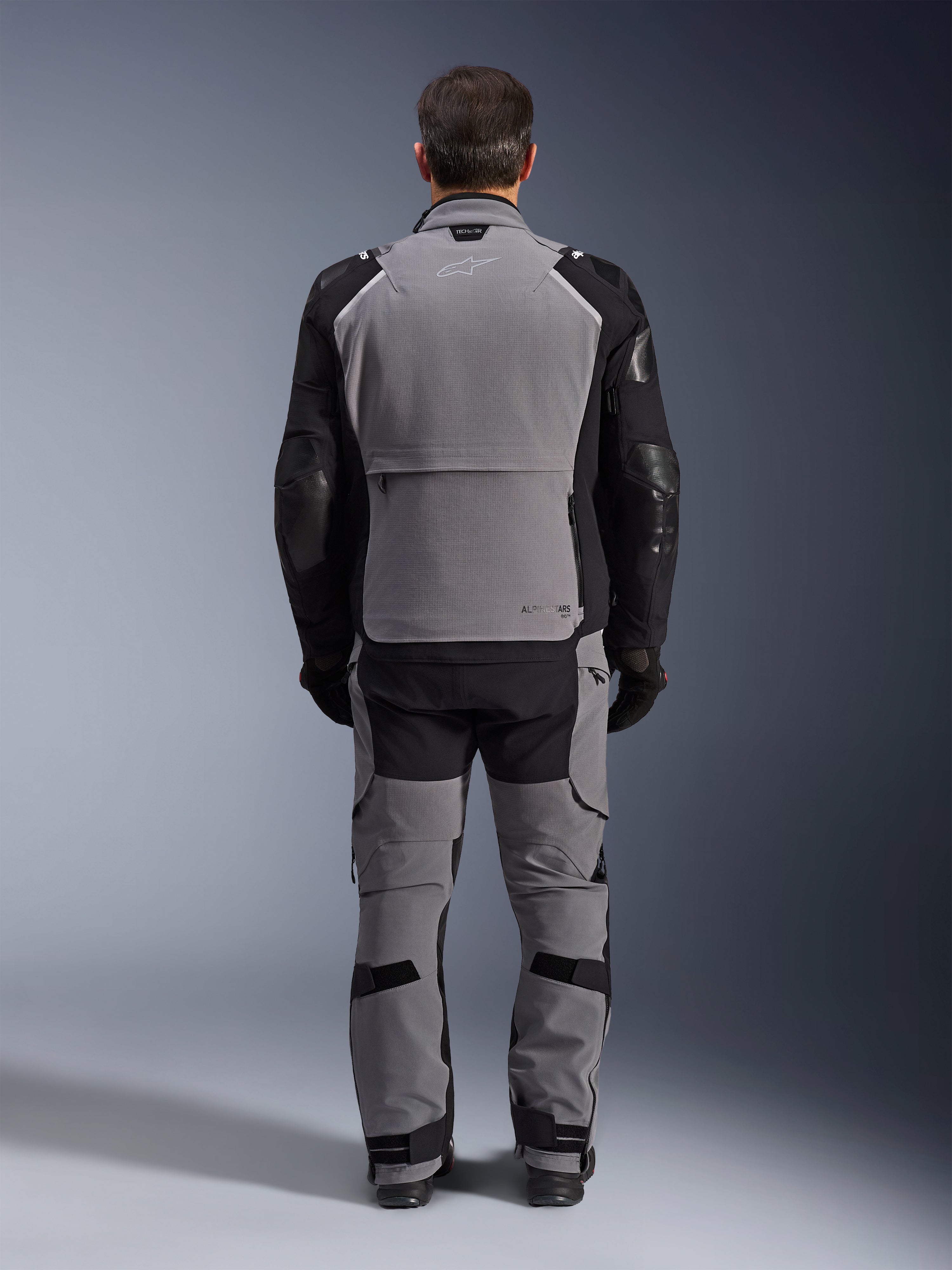 Alpinestars Andes Pro Drystar® XF Jacket, Grigio Scuro Nero, vista posteriore di un pilota che indossa pantaloni coordinati, giacca da moto adventure touring con un ampio pannello posteriore grigio, maniche articolate nere con protezioni e un vano portaoggetti posteriore