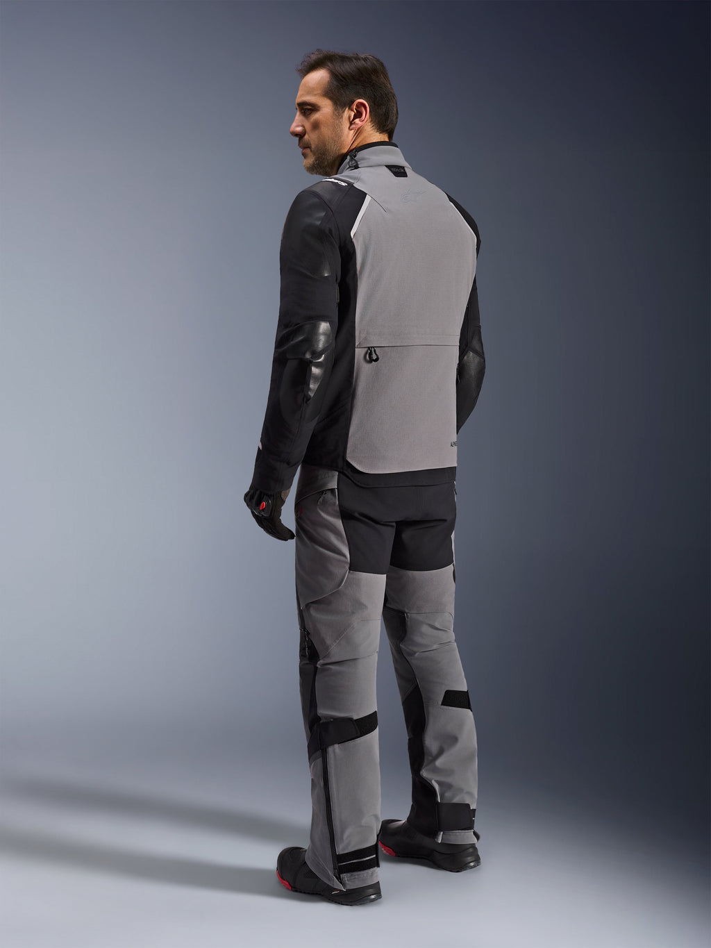 Giacca Alpinestars Andes Pro Drystar® XF, giacca da moto adventure-touring in grigio scuro e nero, vista posteriore su un modello che mostra l'ampio pannello posteriore e le maniche rinforzate, abbinata a pantaloni adventure coordinati, guanti e stivali