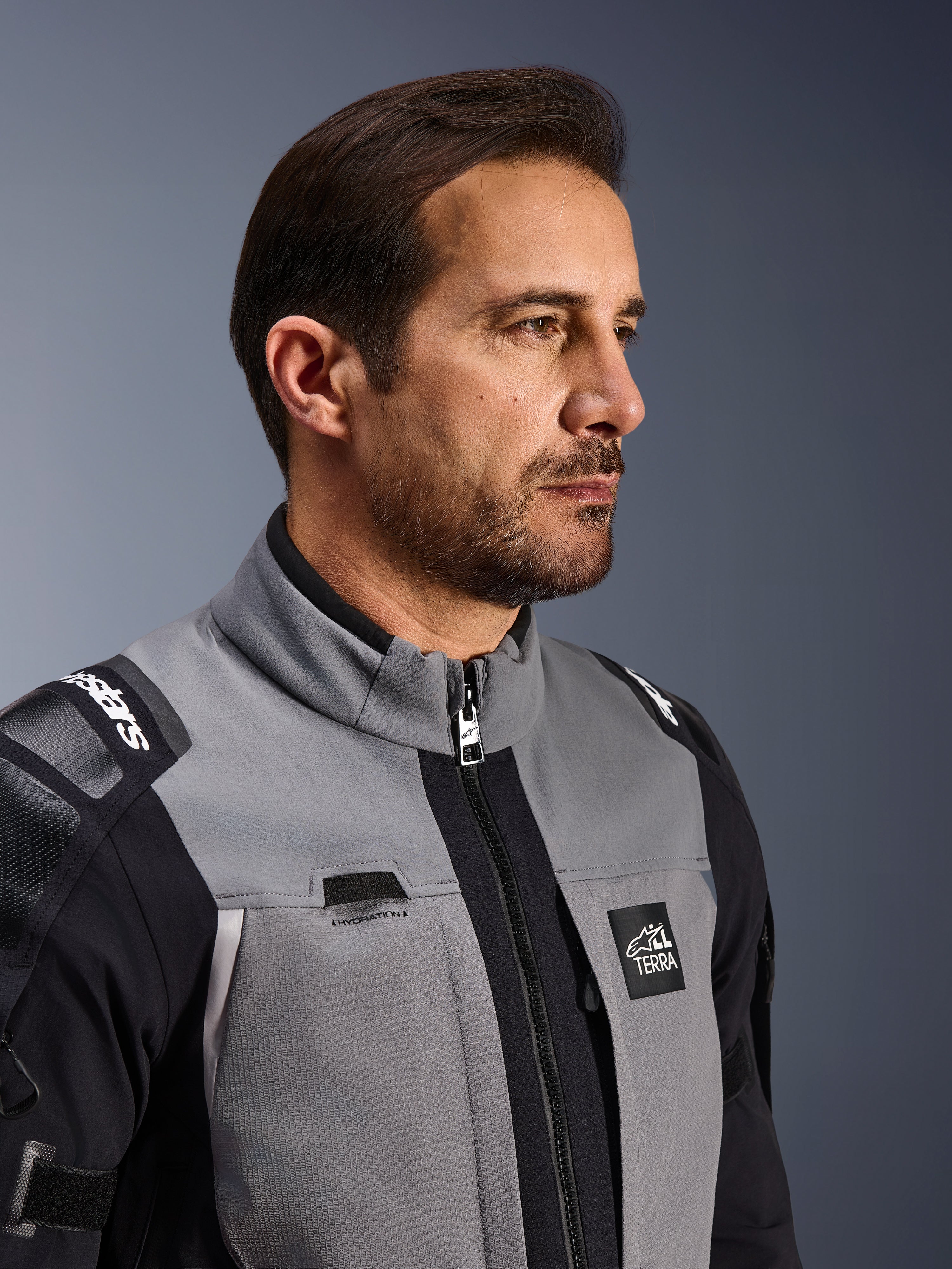 Giacca Alpinestars Andes Pro Drystar® XF, giacca da moto adventure, grigio scuro e nero, primo piano della parte anteriore del busto e del colletto, con logo Terra e dettaglio del passaggio per l'idratazione, indossata da un uomo in uno studio.