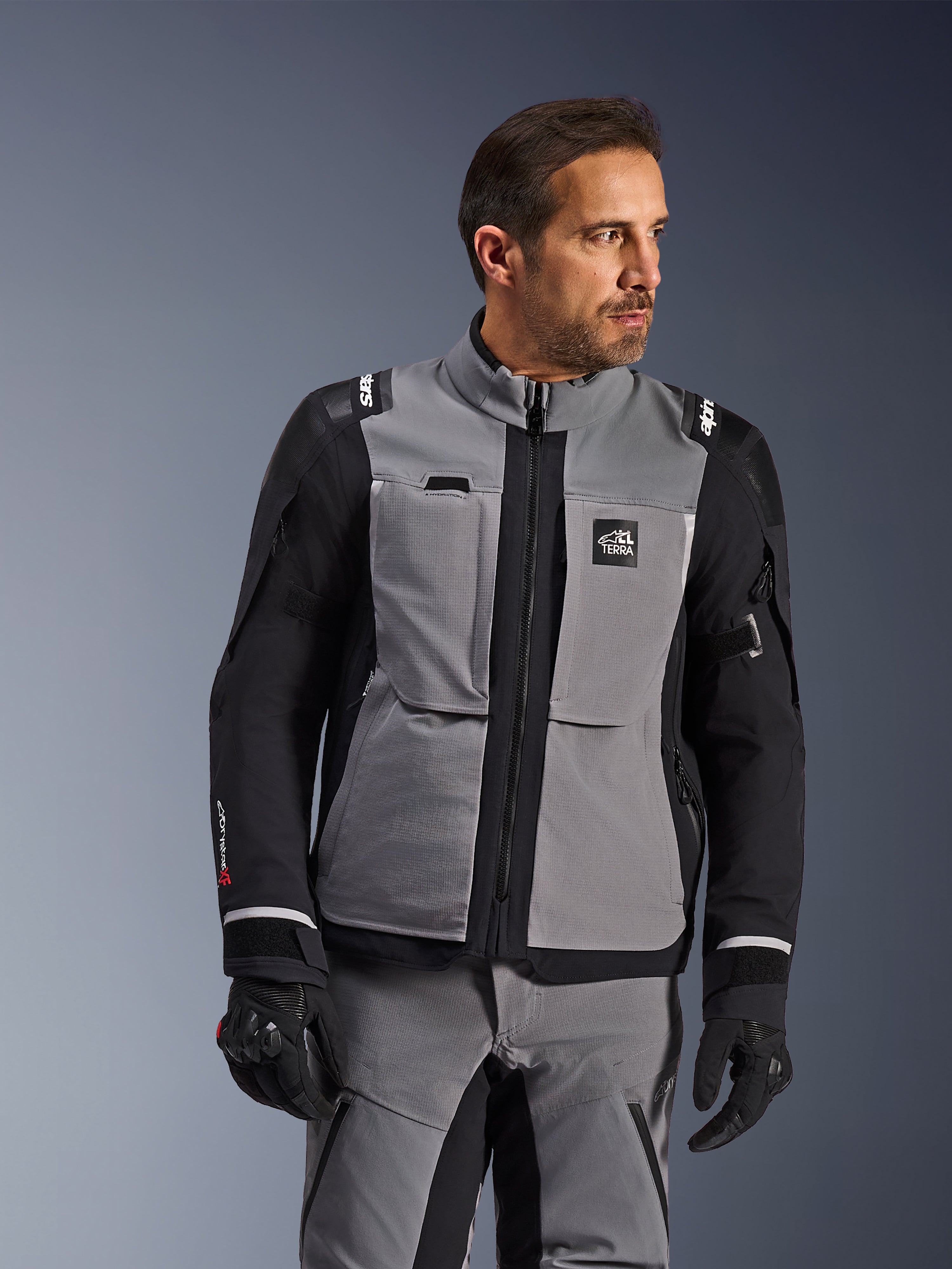 Giacca Alpinestars Andes Pro Drystar® XF in grigio scuro e nero, abbigliamento per guida adventure, indossata da un uomo con pantaloni grigi coordinati e guanti neri, con guscio laminato a 3 strati, tasche utility e logo 'Terra' sul petto