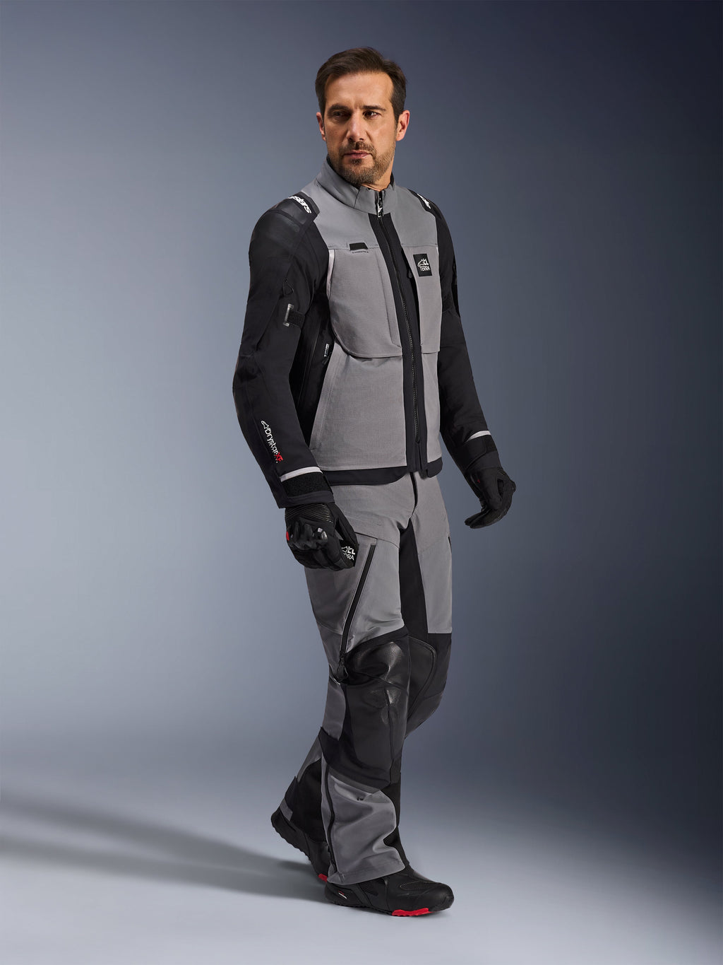 Giacca Alpinestars Andes Pro Drystar® XF, giacca da moto adventure, grigio scuro e nero, indossata da un modello con pantaloni in tessuto coordinati e guanti neri, caratterizzata da un design multitasche, guscio laminato tecnico a 3 strati e vestibilità articolata per viaggi adventure a lunga distanza.