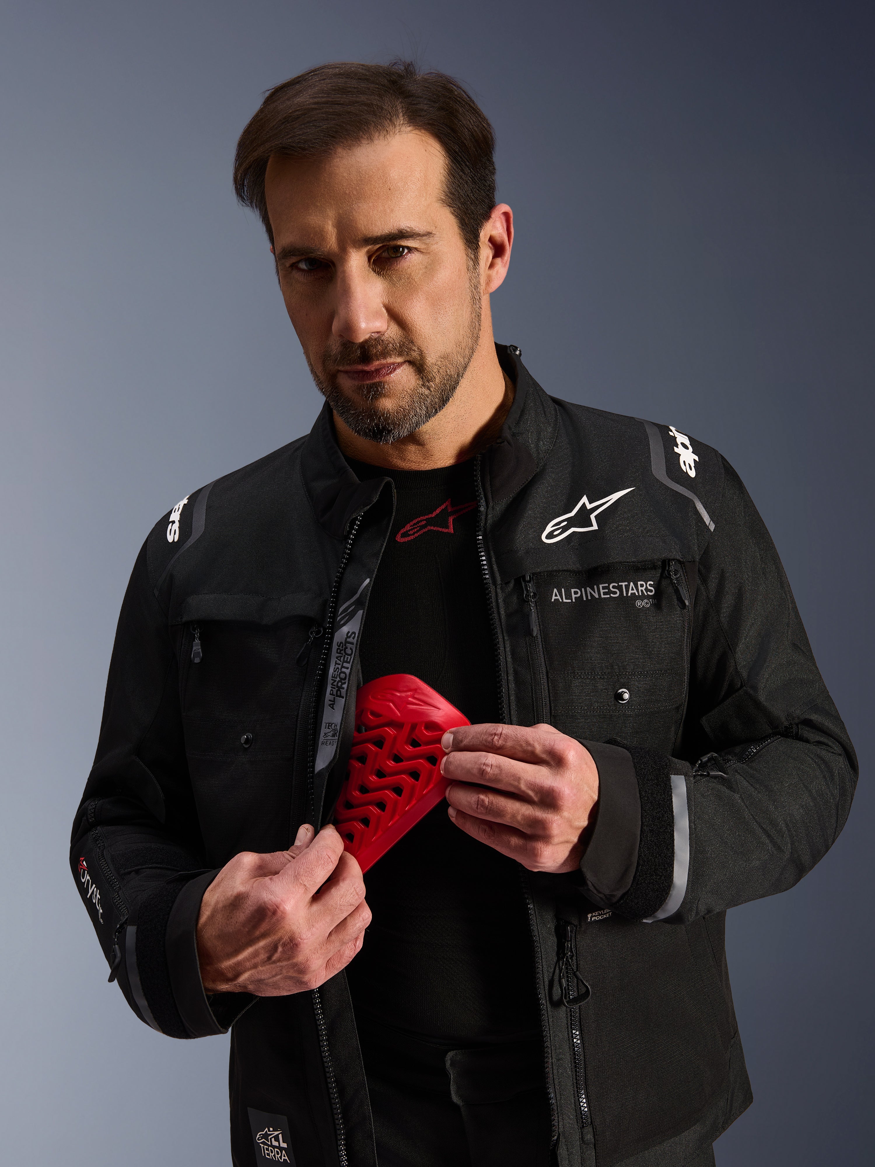 Giacca Alpinestars Cusco Drystar® nera, modello maschile con una protezione Nucleon Flex Pro rossa, giacca da moto adventure touring con branding bianco, costruzione in tessuto tecnico con cerniere di ventilazione e tasche multiple