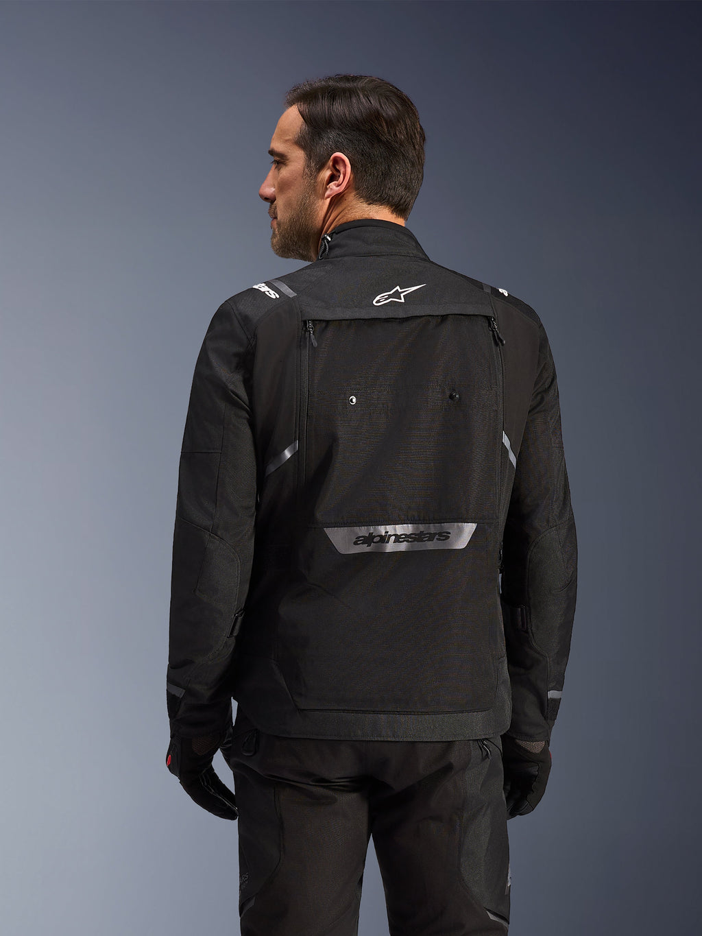 Pilota visto da dietro che indossa la giacca Alpinestars Cusco Drystar® in nero, giacca in tessuto adventure touring con ampia tasca posteriore e loghi riflettenti, abbinata a pantaloni e guanti neri coordinati