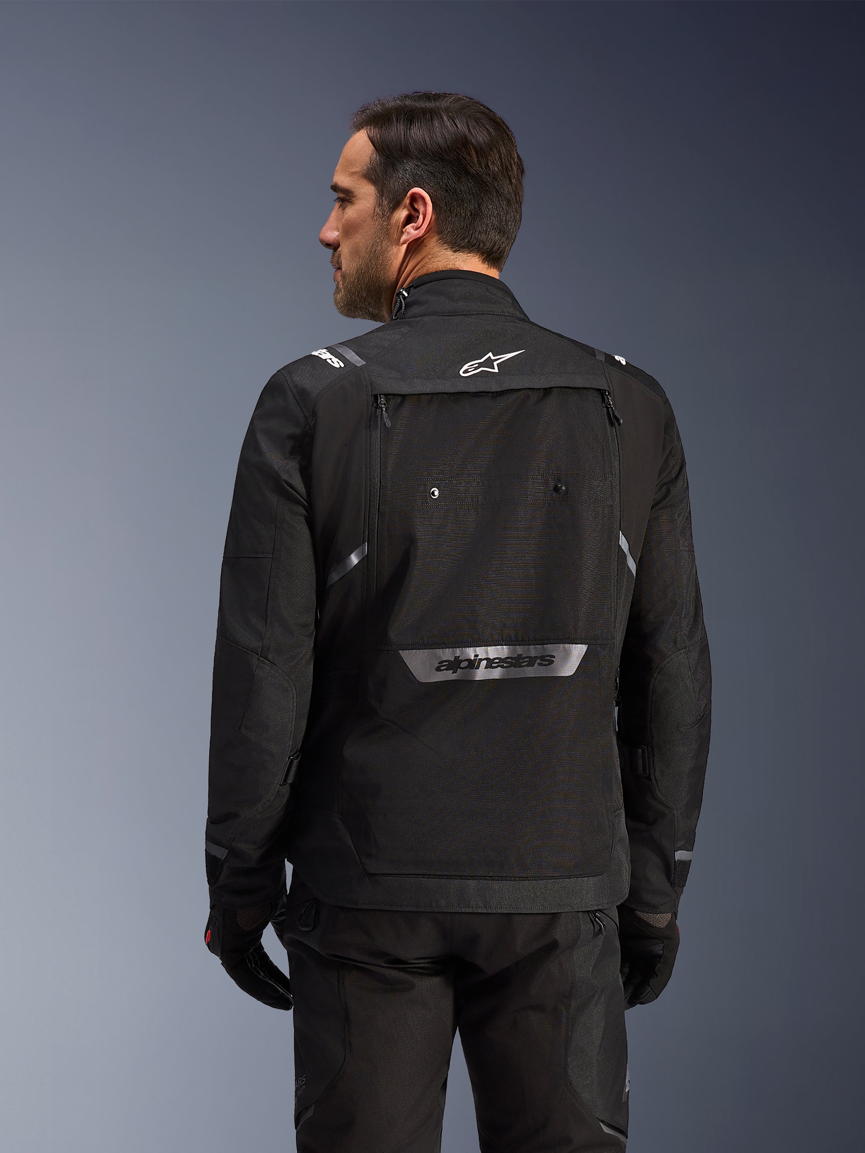 Pilota visto da dietro che indossa la giacca Alpinestars Cusco Drystar® in nero, giacca in tessuto adventure touring con ampia tasca posteriore e loghi riflettenti, abbinata a pantaloni e guanti neri coordinati
