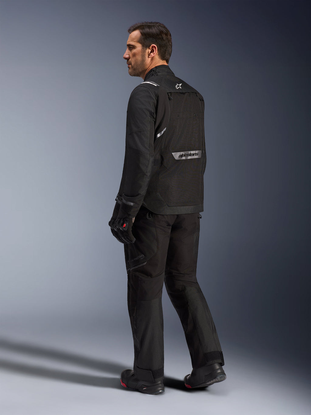 Giacca Alpinestars Cusco Drystar®, giacca da moto nera, vista posteriore di un modello maschile con equipaggiamento completo, con branding riflettente sulla schiena, costruzione in tessuto con pannelli tecnici, outfit coordinato per road adventure touring