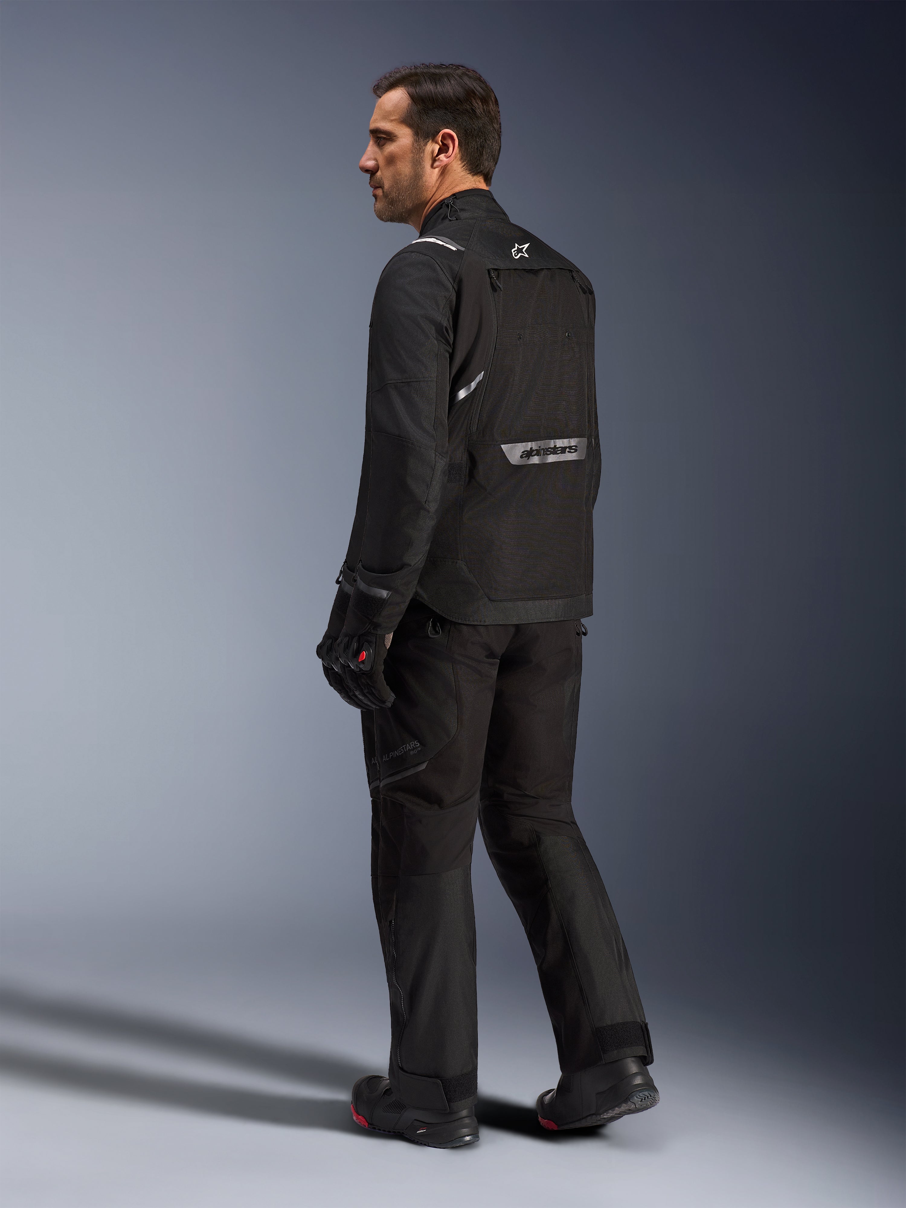 Giacca Alpinestars Cusco Drystar®, giacca da moto nera, vista posteriore di un modello maschile con equipaggiamento completo, con branding riflettente sulla schiena, costruzione in tessuto con pannelli tecnici, outfit coordinato per road adventure touring