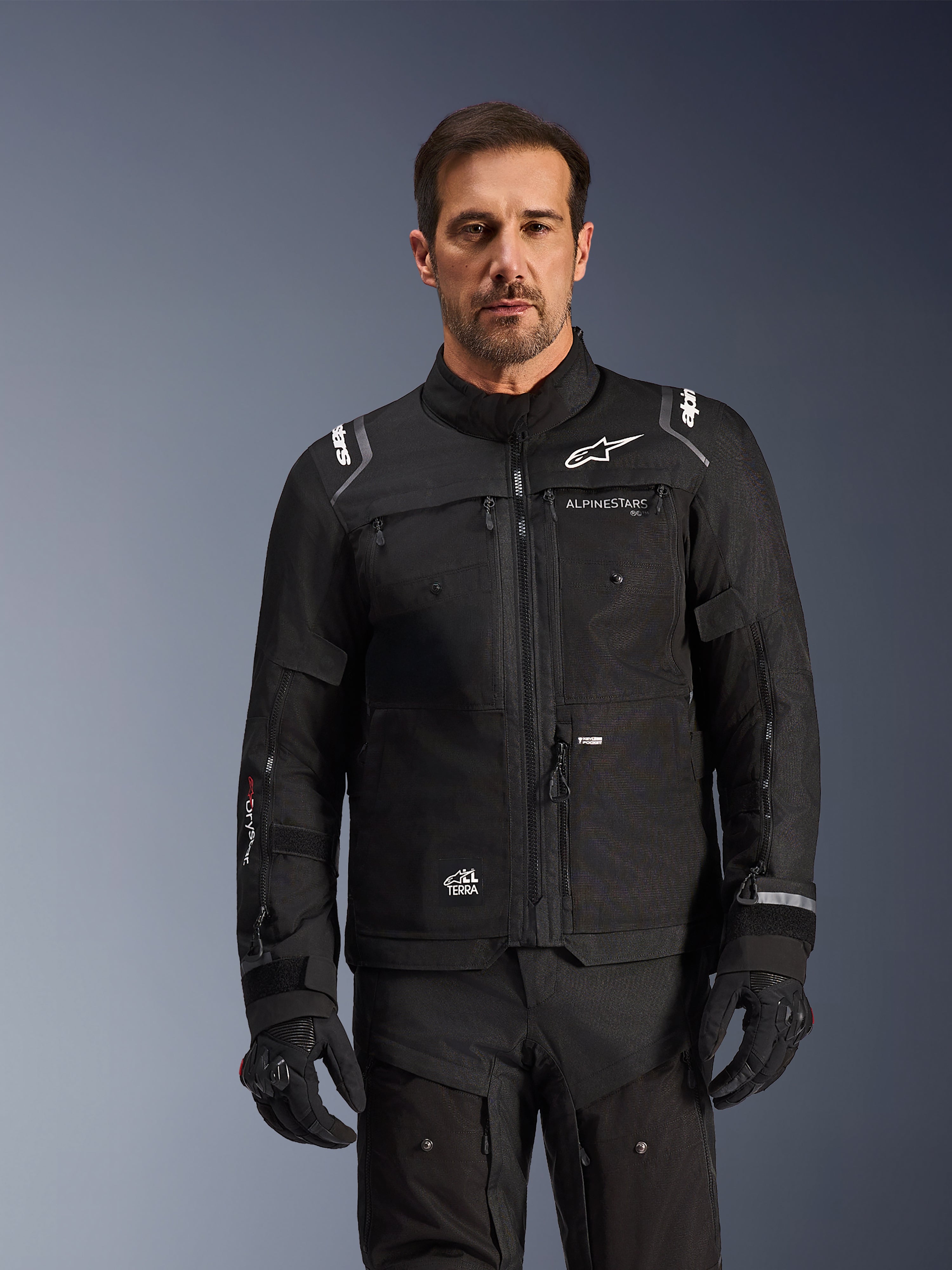 Giacca Alpinestars Cusco Drystar®, giacca da moto nera, indossata da un modello con guanti e pantaloni neri coordinati, caratterizzata da una costruzione in tessuto tecnico con loghi Alpinestars bianchi, progettata per l'adventure touring su strada