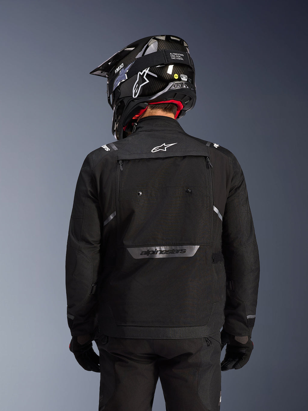 Giacca Alpinestars Cusco Drystar®, nero, vista posteriore di un pilota con casco nero e guanti, presenta ampi pannelli di ventilazione, logo bianco sul collo e marchio riflettente sulla parte inferiore della schiena, abbigliamento adventure touring